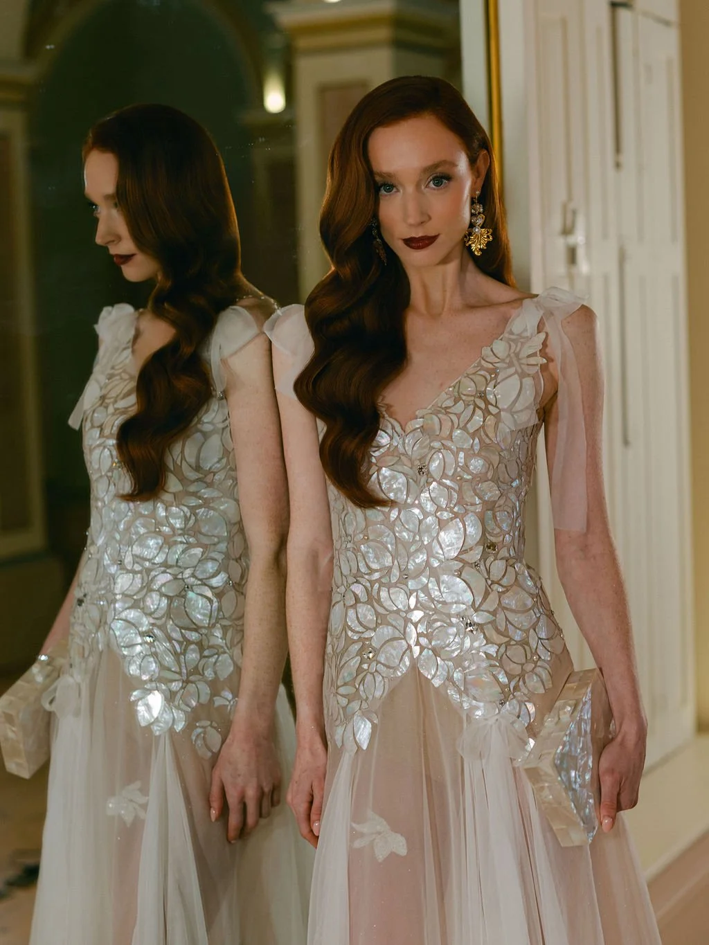 A woman with long, wavy auburn hair and dark lipstick standing beside a mirror, wearing a white dress with floral applique and holding a clutch.
