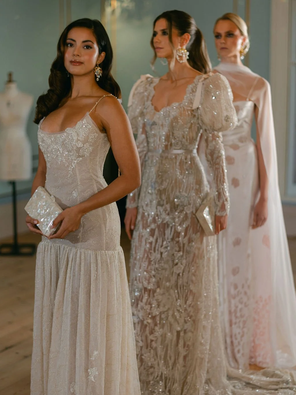 Four women in elegant, ornate formal dresses standing indoors, with the woman in front holding a clutch purse.