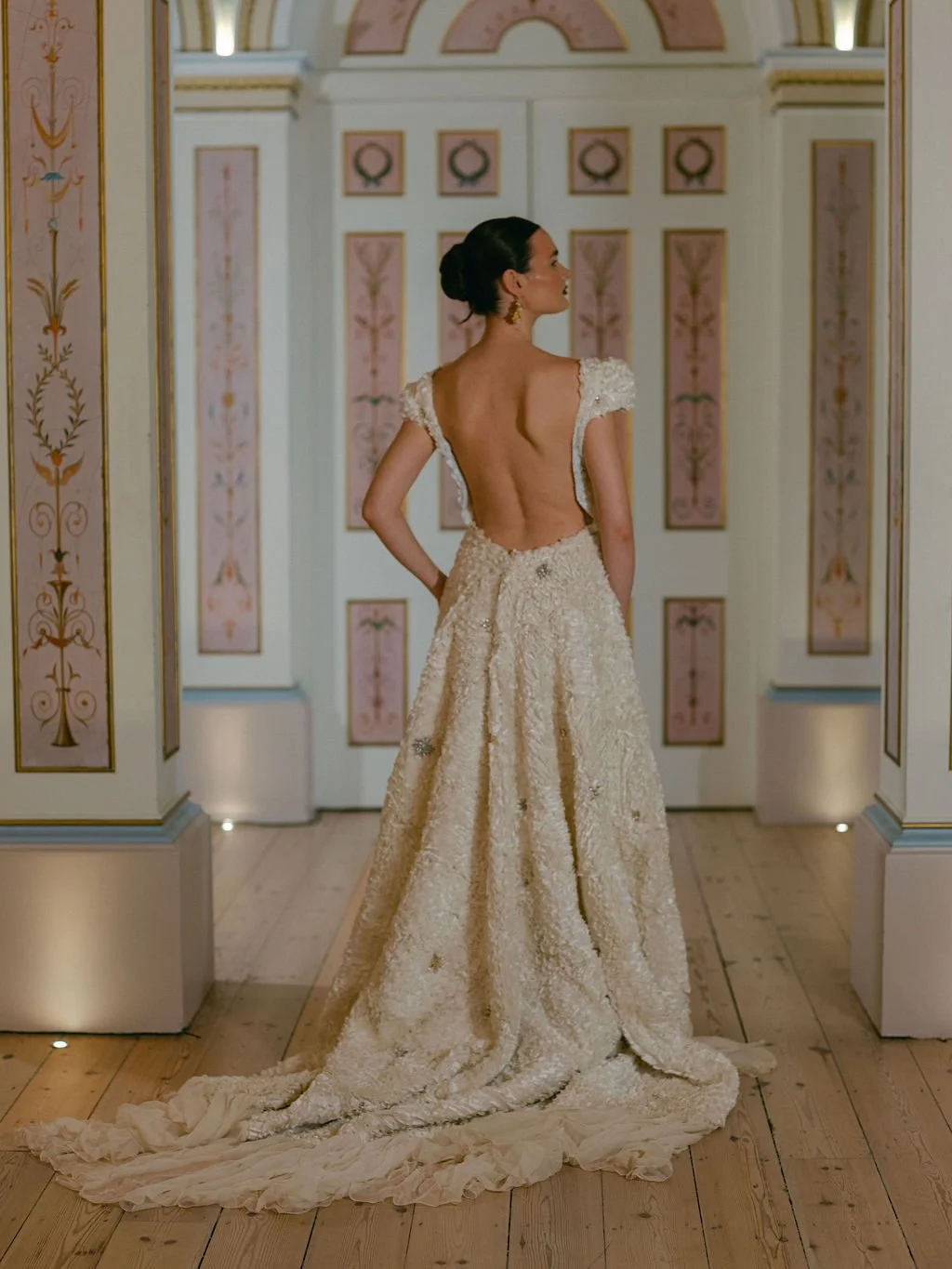 Woman in an elegant, backless wedding gown with floral details standing in a decorated, pastel-colored room.