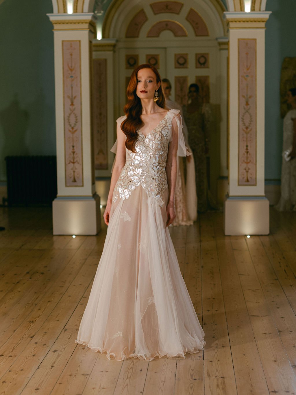 A woman in an elegant gown displays a floral design with layered sheer fabric on the shoulders, standing in a historically decorated room with a wooden floor and painted archway.