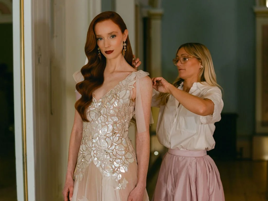 A woman with long red hair in an elegant white dress is having her dress adjusted by another woman with blond hair and glasses, in a cream blouse and pink skirt, in a room with a mirror and chandelier.