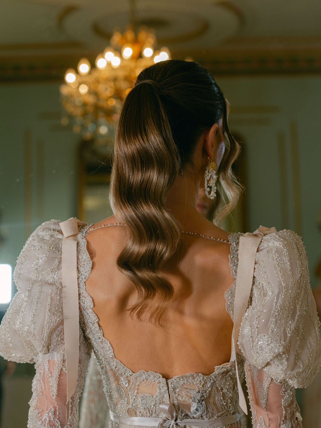 Back of a woman with long wavy hair, wearing a vintage wedding dress with lace details and puffed sleeves, standing in a grand, elaborately decorated room.