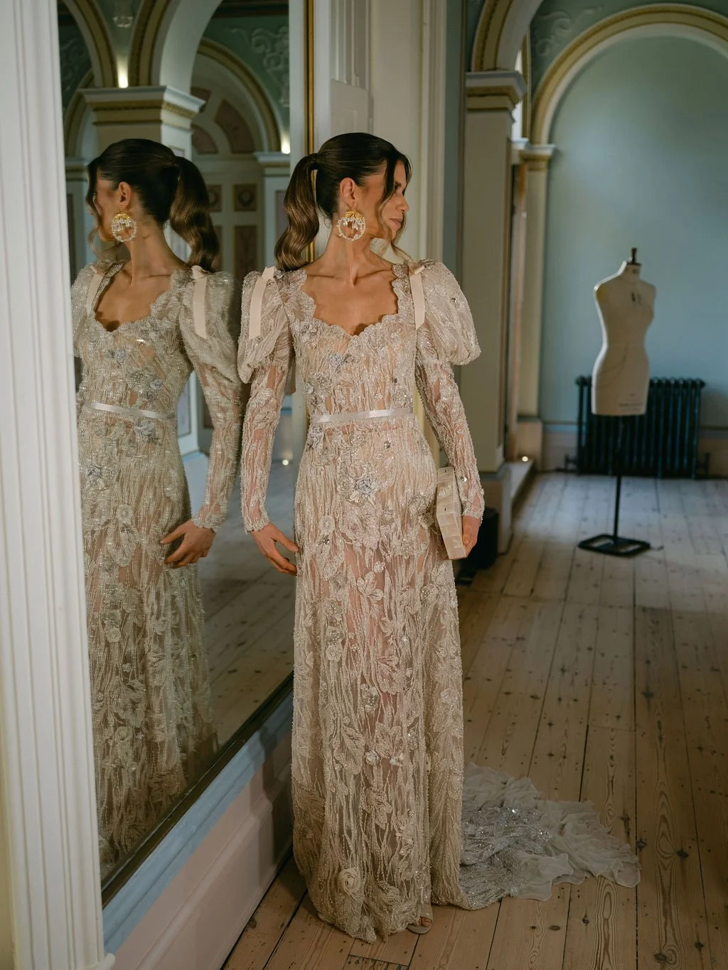 Woman looking at her reflection in a mirror, wearing an elegant, long, lace dress with floral embroidery and puffed sleeves, in a room with high ceilings, arched walls, and a mannequin in the background.
