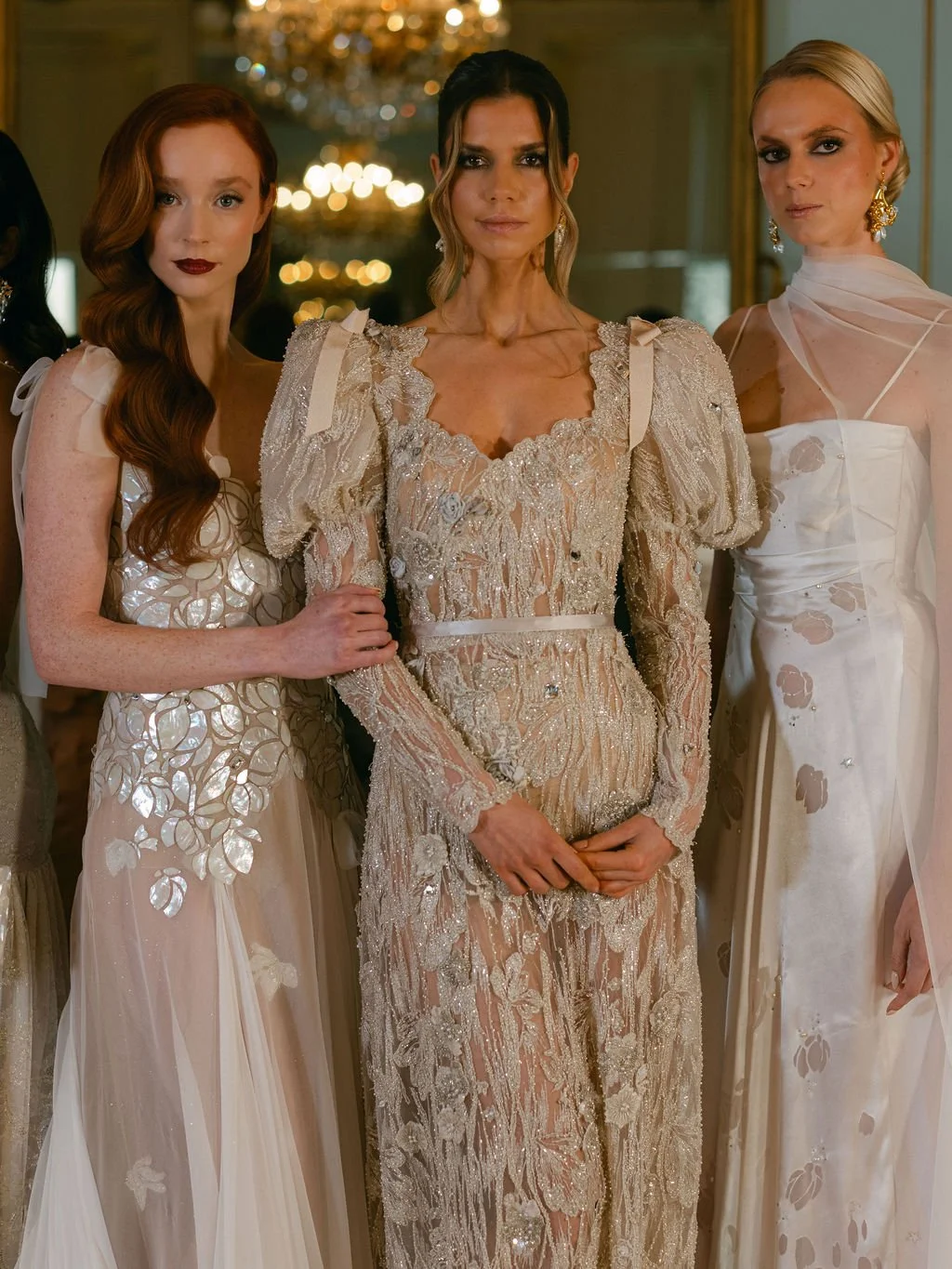 Three women in elegant, ornate dresses at a formal event in a lavish setting with a chandelier in the background.