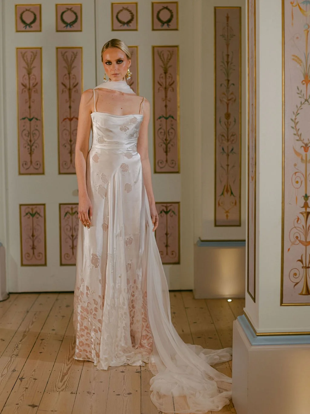 A woman in an elegant, satin and lace wedding dress with a long train and spaghetti straps, standing in a decorated room with cream and pink patterned walls.
