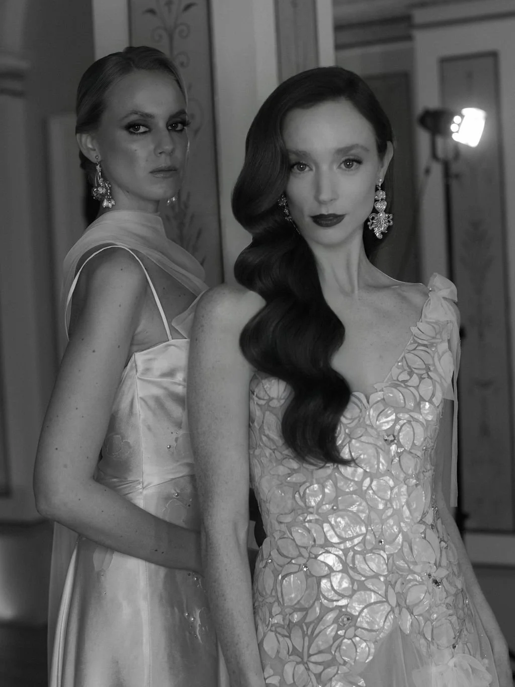 Two women in elegant dresses and jewelry look into the camera; one has dark hair styled in waves, and the other has light hair styled back. They are indoors with a mirror and a light visible in the background.