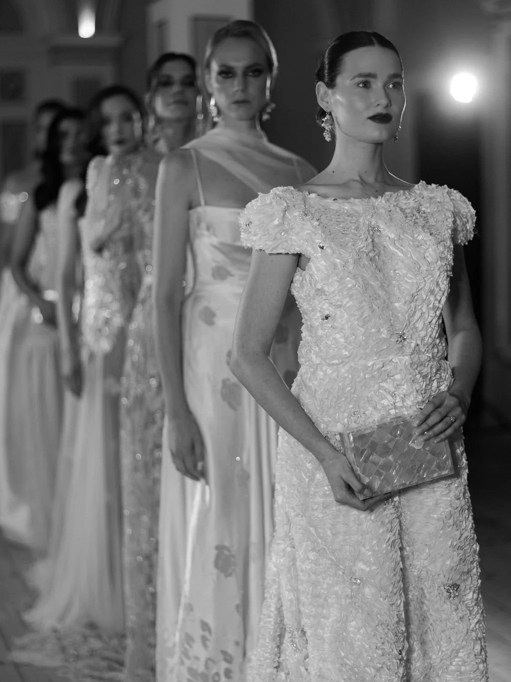 Line of elegantly dressed women in formal gowns, standing in a dimly lit room, with one woman in the foreground holding a clutch purse.