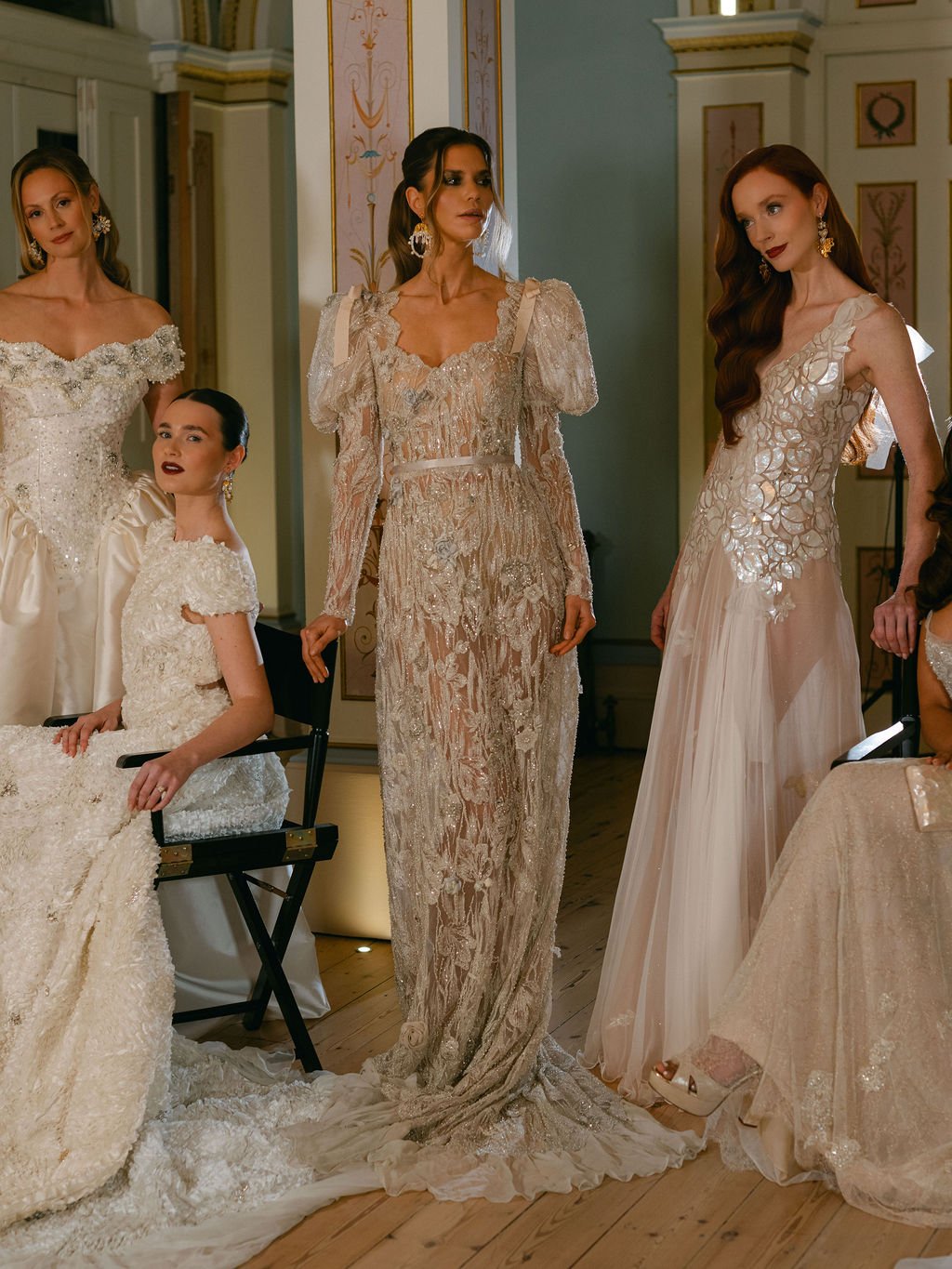 Four women in elegant wedding dresses inside a decorated room, some seated and some standing, with intricate details and a classical interior design.