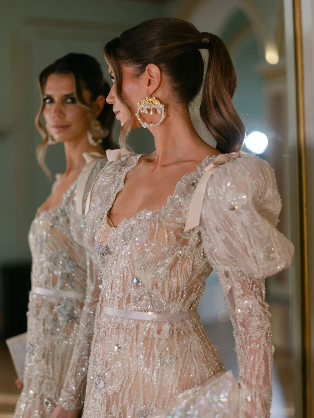 Woman in an ornate, shiny, white gown with puffed sleeves, standing in front of a mirror, wearing large, decorative earrings and looking at her reflection.