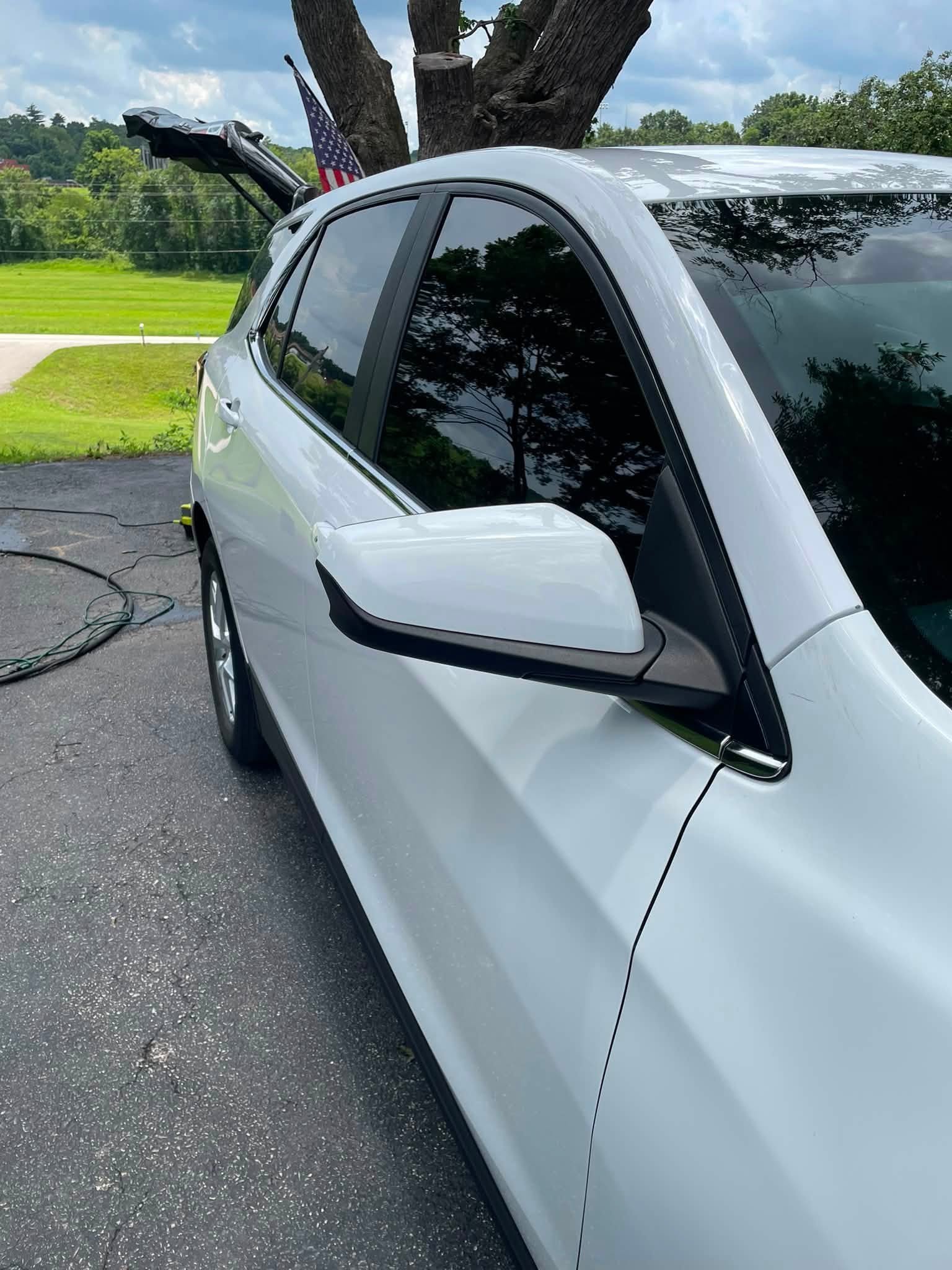 A silver electric vehicle parked outdoors on a paved surface, with a large tree in the background and two American flags attached to the car's rear. The rear trunk is open, and charging cables are connected to the vehicle.