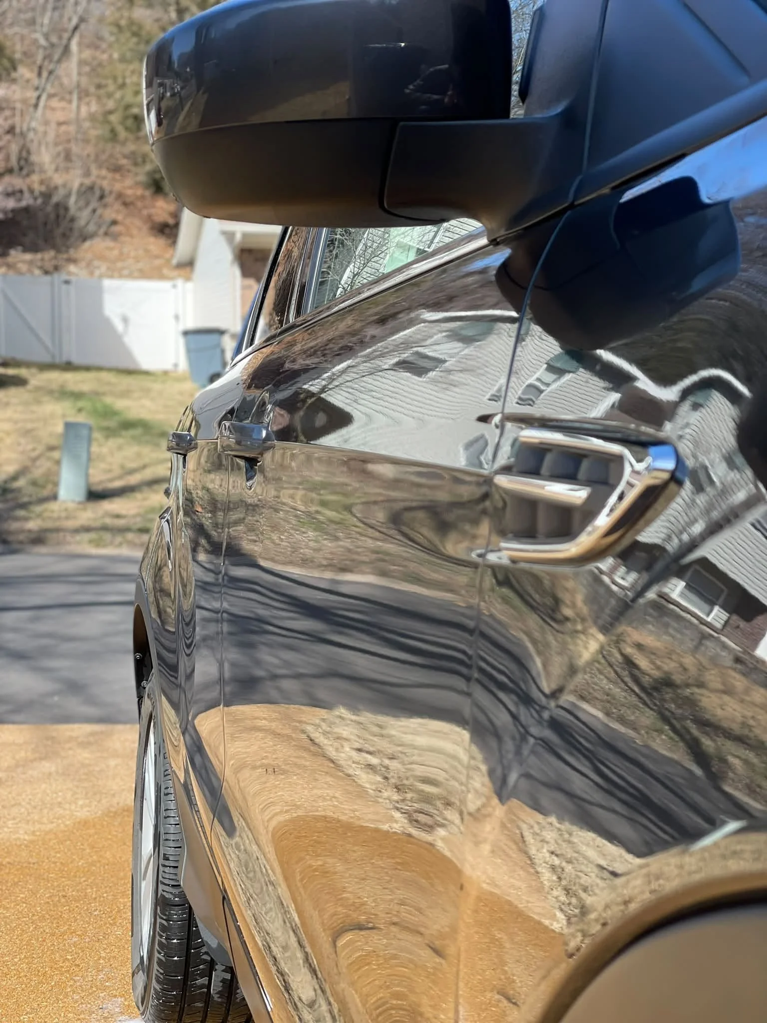 Close-up of a shiny black car's side, with a black side mirror and a window, reflecting a suburban yard with grass, trees, a white fence, and a house in the background. The photo is taken outdoors during daytime.