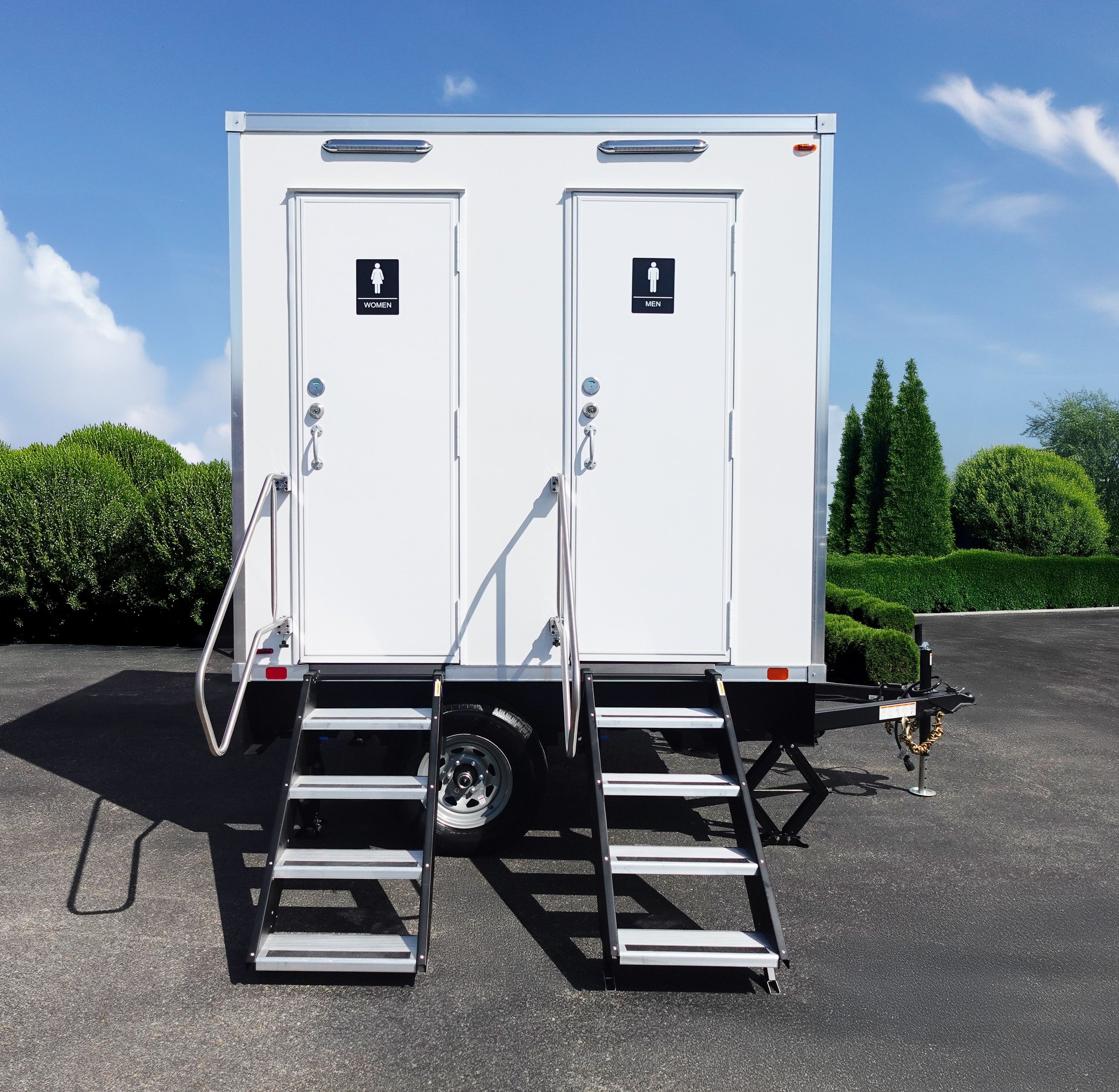 A white mobile restroom trailer with separate doors for women and men, featuring black signs with white icons and text. The trailer has steps leading up to the entrances, silver handles, and is set against a background of trees, bushes, and a partly cloudy sky.