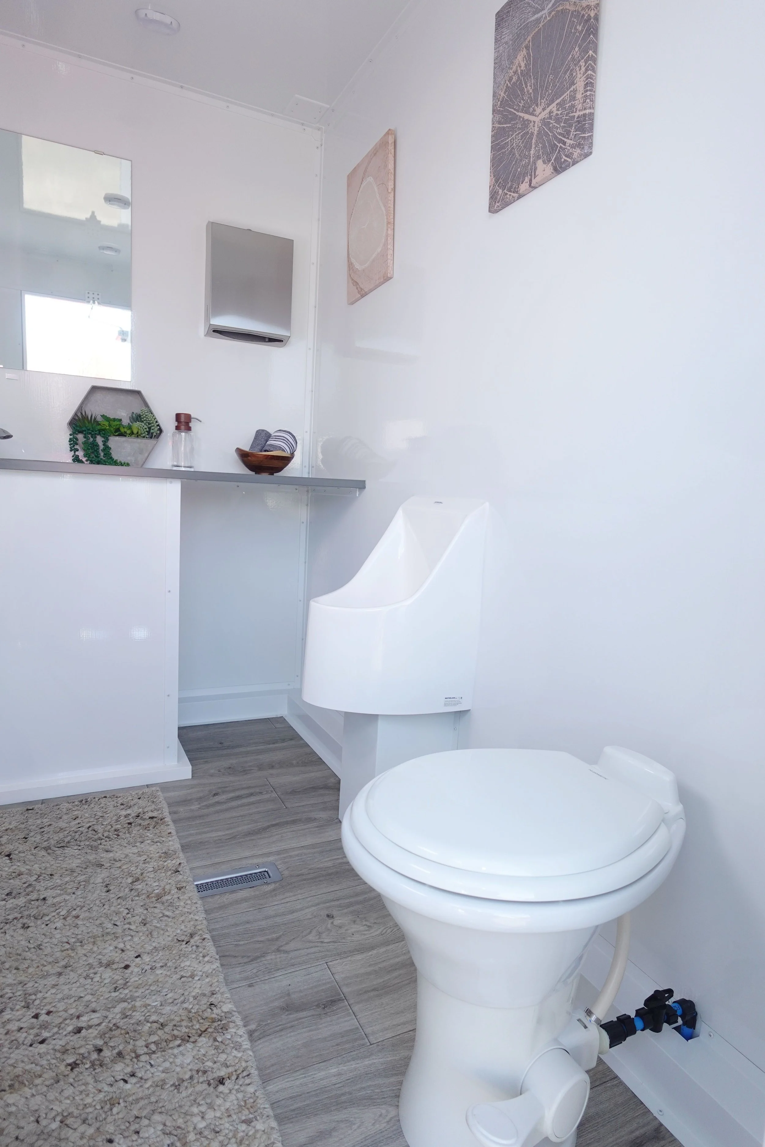 A modern bathroom featuring a white toilet, a white vanity, a wall mirror, decorative art pieces, a small shelf with plants and towels, a wall-mounted hand dryer, a bathroom mat, and wooden-look flooring.