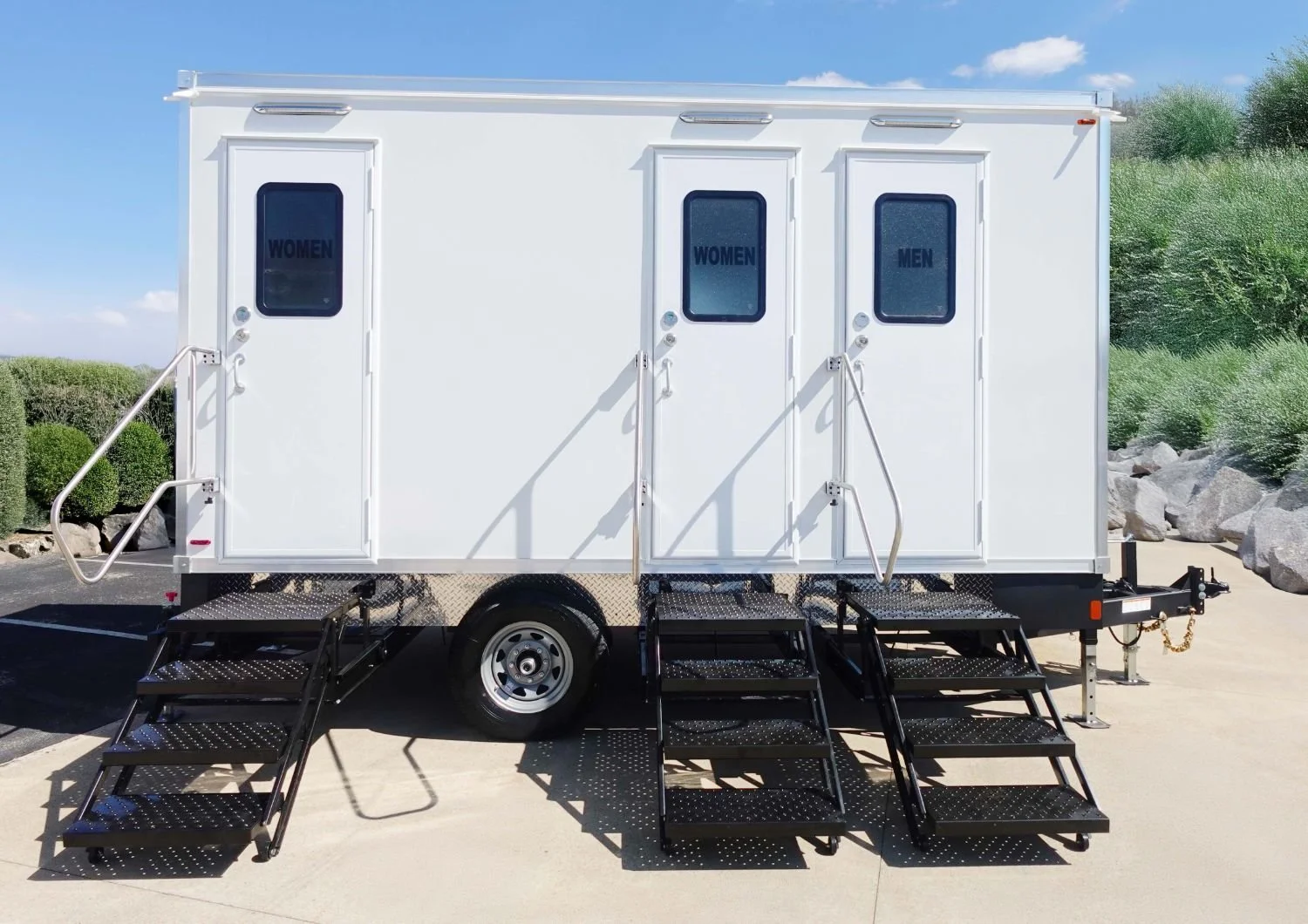 White portable restroom trailer with separate entrances for women and men, featuring black signs on the doors, with outdoor stairs and a black metal ramp for accessibility, set against a clear sky and greenery.