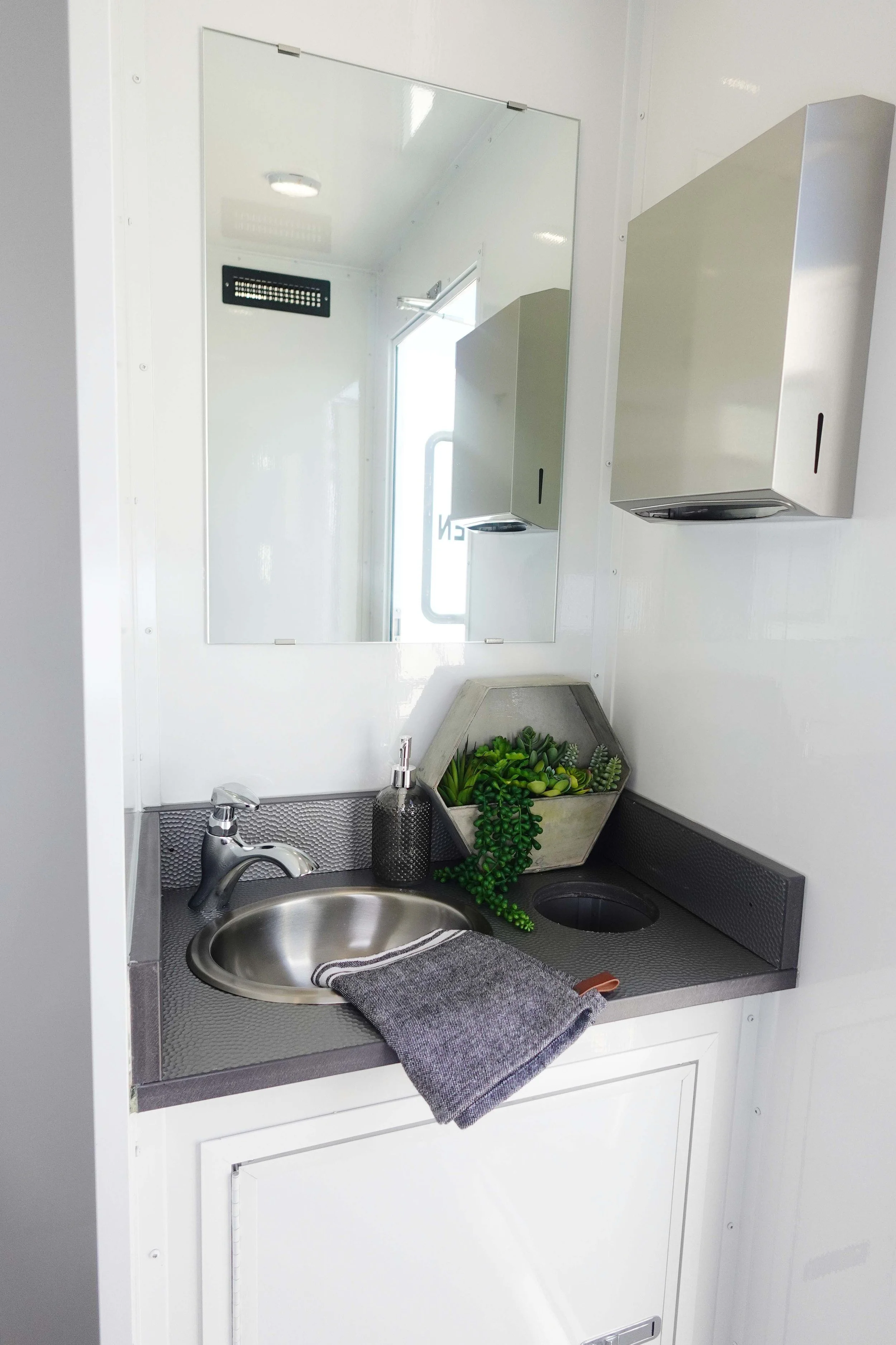 Small kitchen sink with a silver faucet, gray towel, soap dispenser, and a small plant in a hexagonal container, with a mirror above the sink and metal cabinets on the wall.