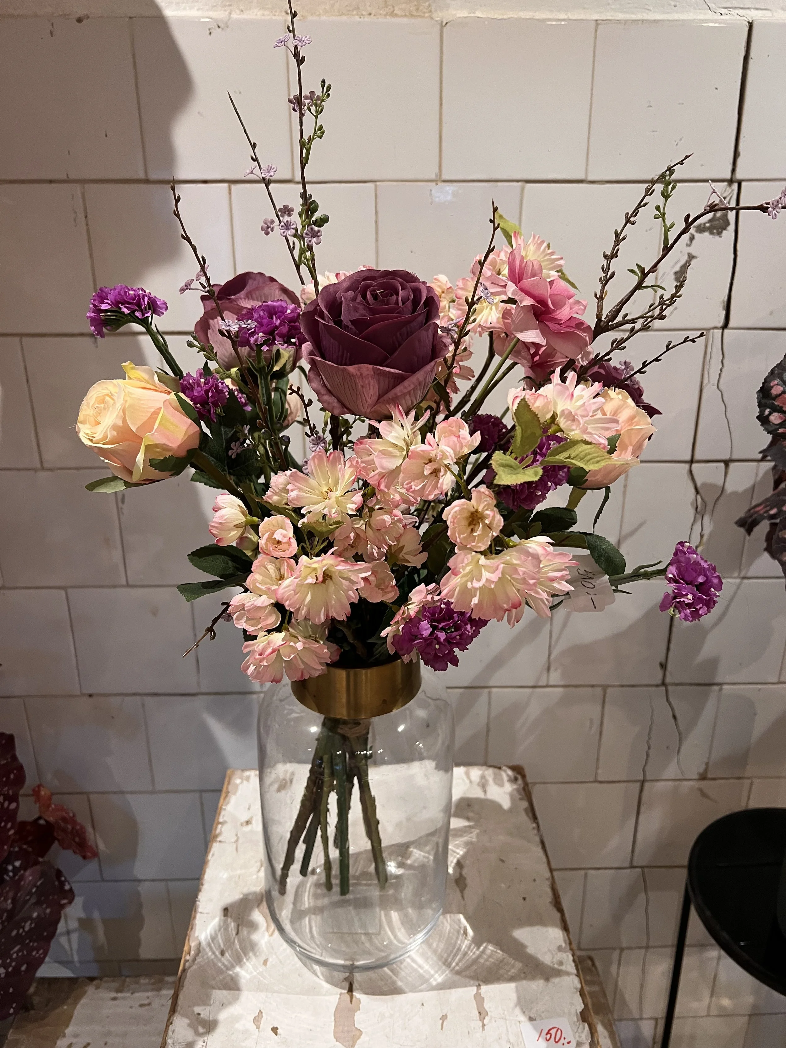 En bukett av blommor i en glasvasj på ett vitt träbord. Blommorna inkluderar mörk lila ros, ljusrosa ros, lila och rosa små blommor samt små grenar med knoppar. Bakgrunden är vita glaserade tegelstenar.