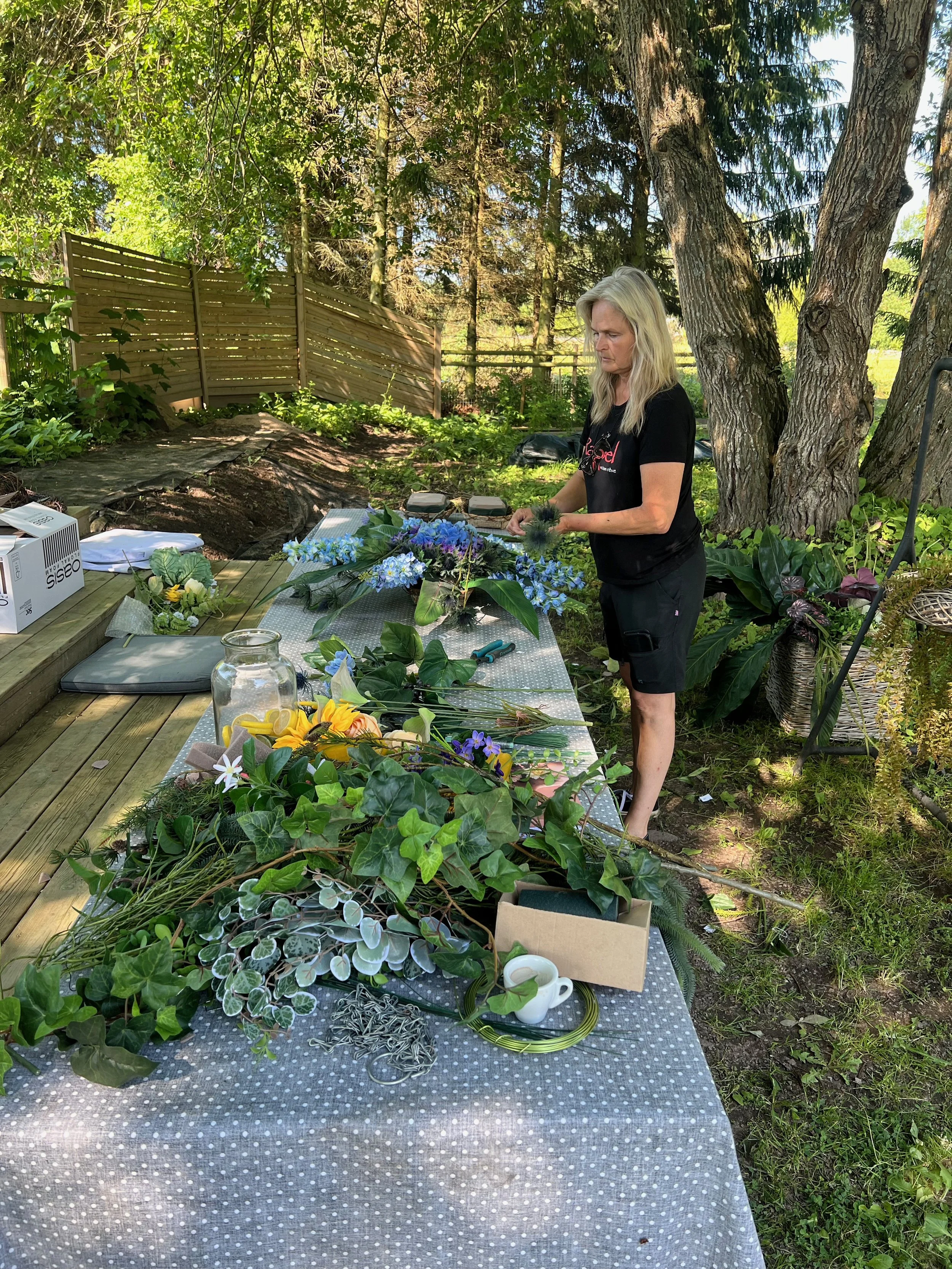 En kvinna med långt blond hår står vid ett bord med olika växter, blommor och träd i bakgrunden. Utomhus i en trädgård under dagtid.