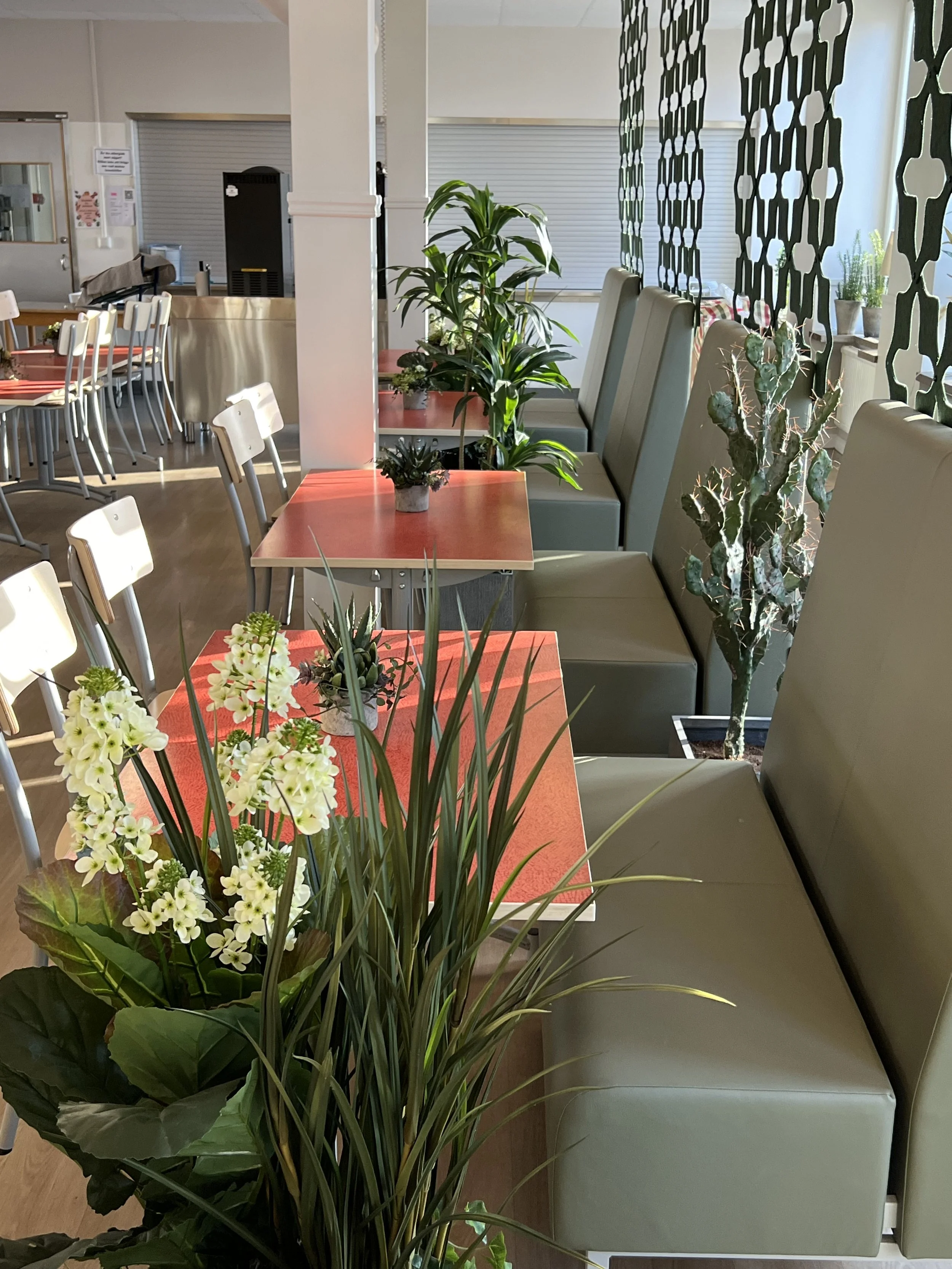Inomhusmiljö med gröna växter, matbord med blommor, gråa sittbänkar, mittemot fönster med utsikt, och en vägg av dekorativa gröna mönster. Fönstret har ett fönsterbräde med fler växter.
