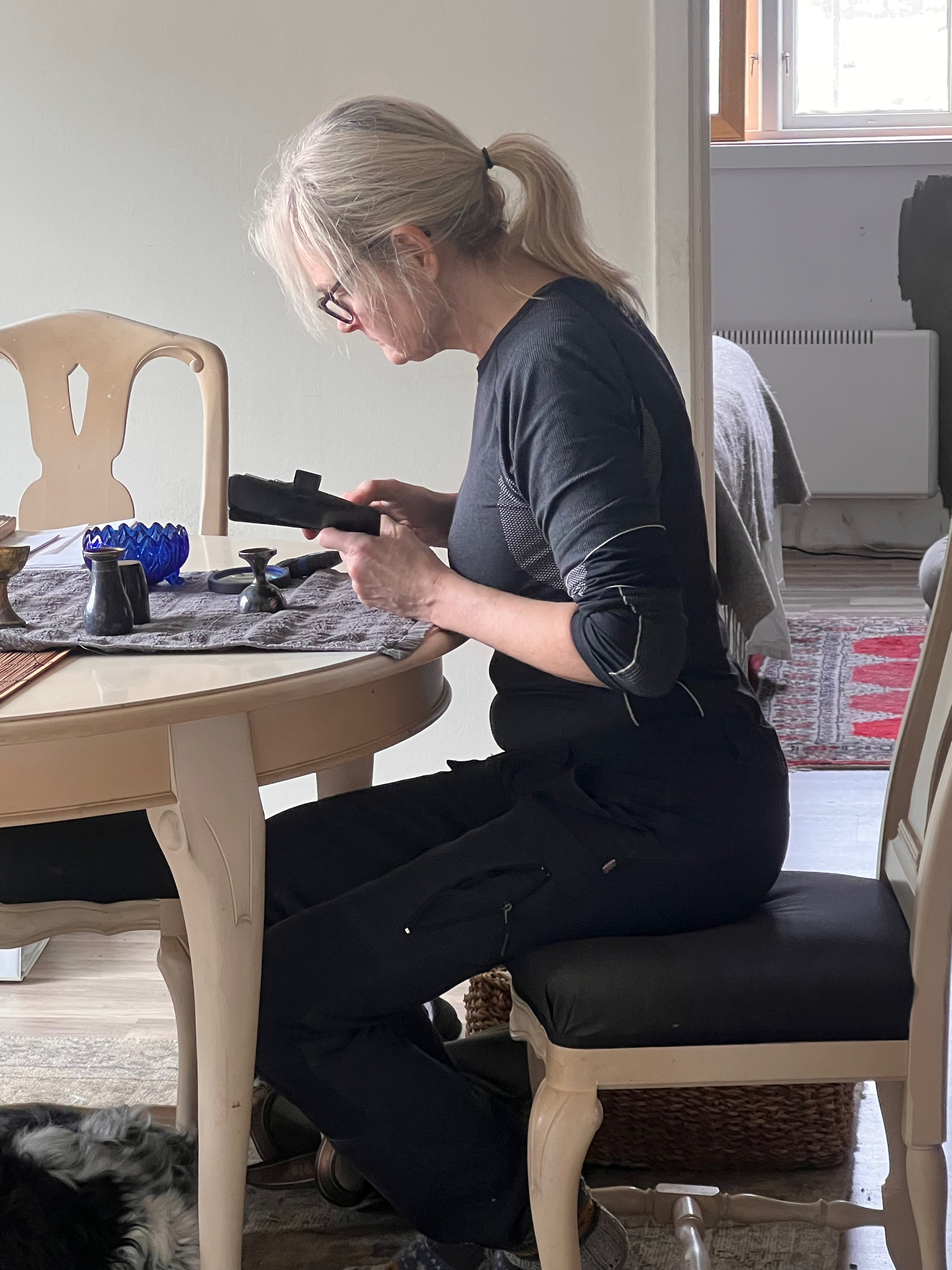 Kvinna med blont hår i hästsvans, glasögon, klädd i svart, sitter på en stol vid ett bord och tittar på sin telefon. Bakom henne ett fönster, del av ett annat rum med matta och soffor.