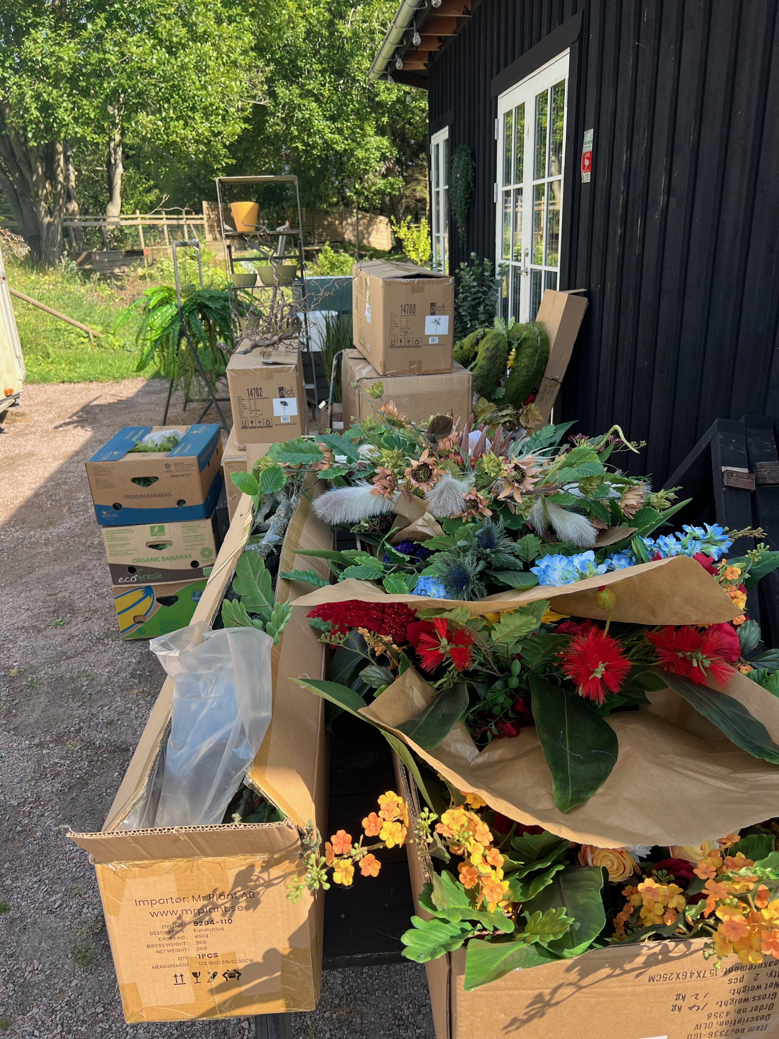 En uteplats med kartonger och blomsterarrangemang utomhus, nära ett svart hus med vita fönster. Det finns gröna träd och buskar i bakgrunden.