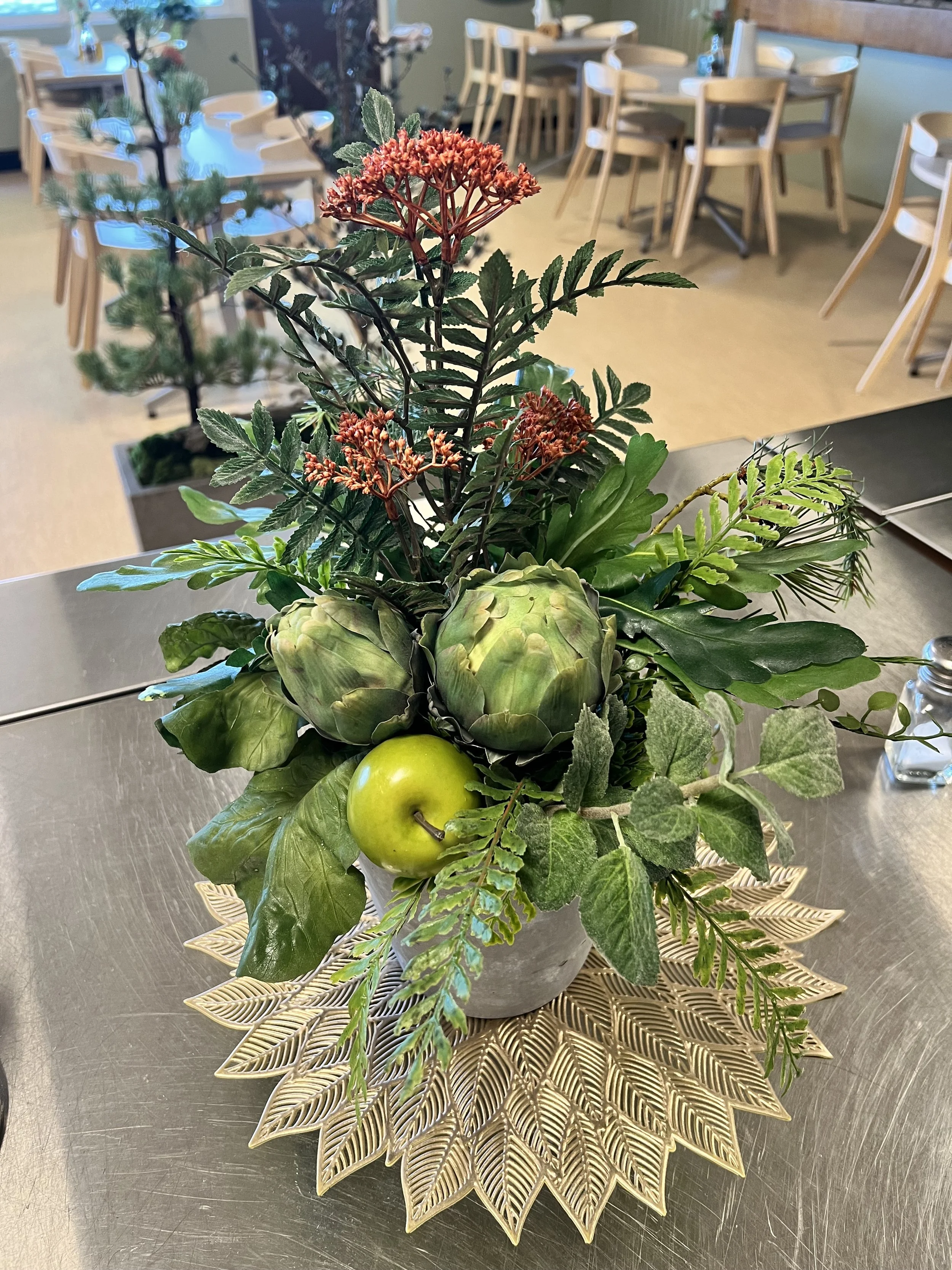 Blomsterarrangemang med gröna blad, ett äpple och en konstgjord blomma i en kruka, placerad på en guldfärgad metallbricka.