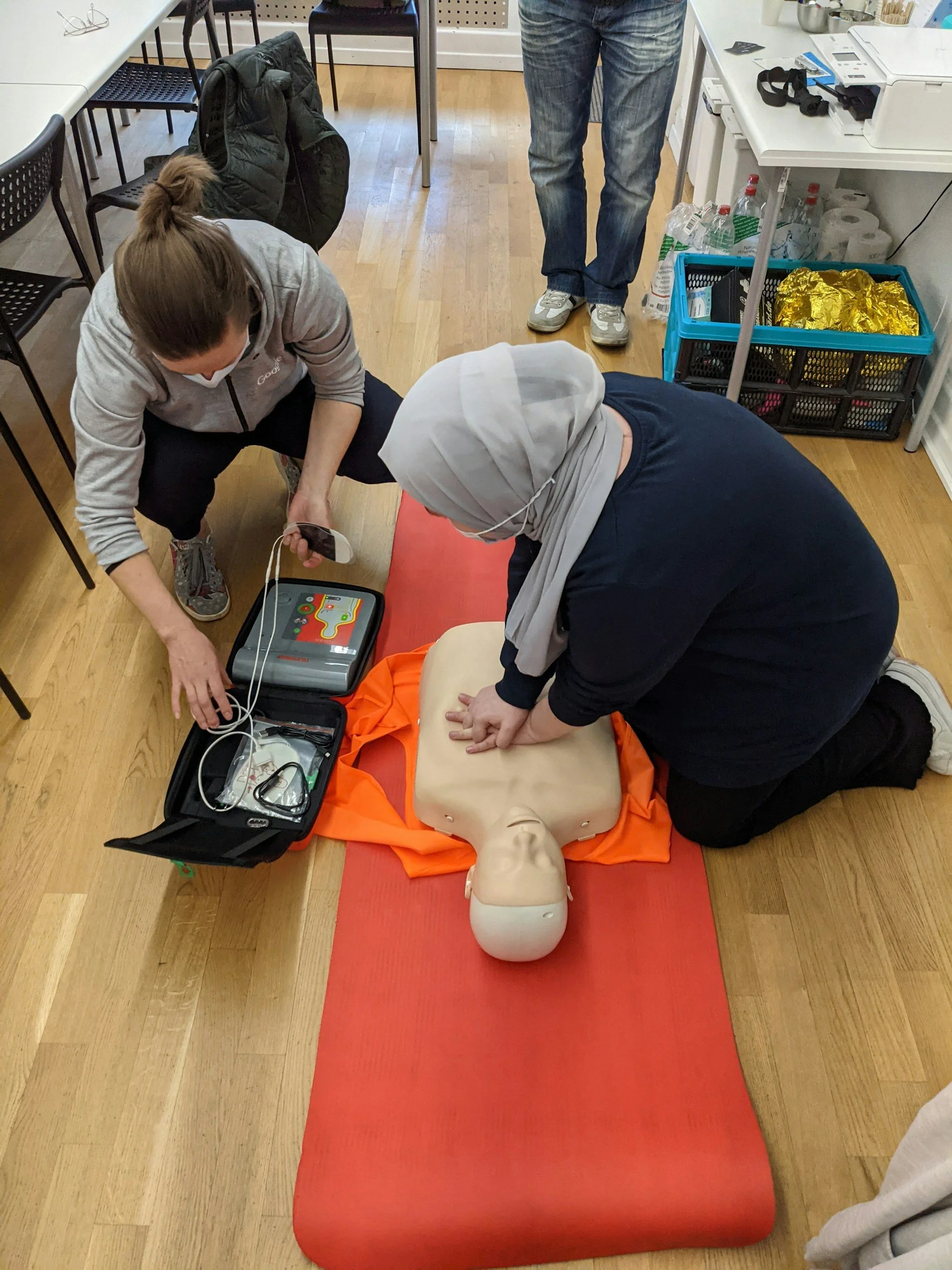 Two women performing CPR on a mannequin in a training room. One woman is kneeling, and the other is leaning over the mannequin, with a defibrillator nearby.