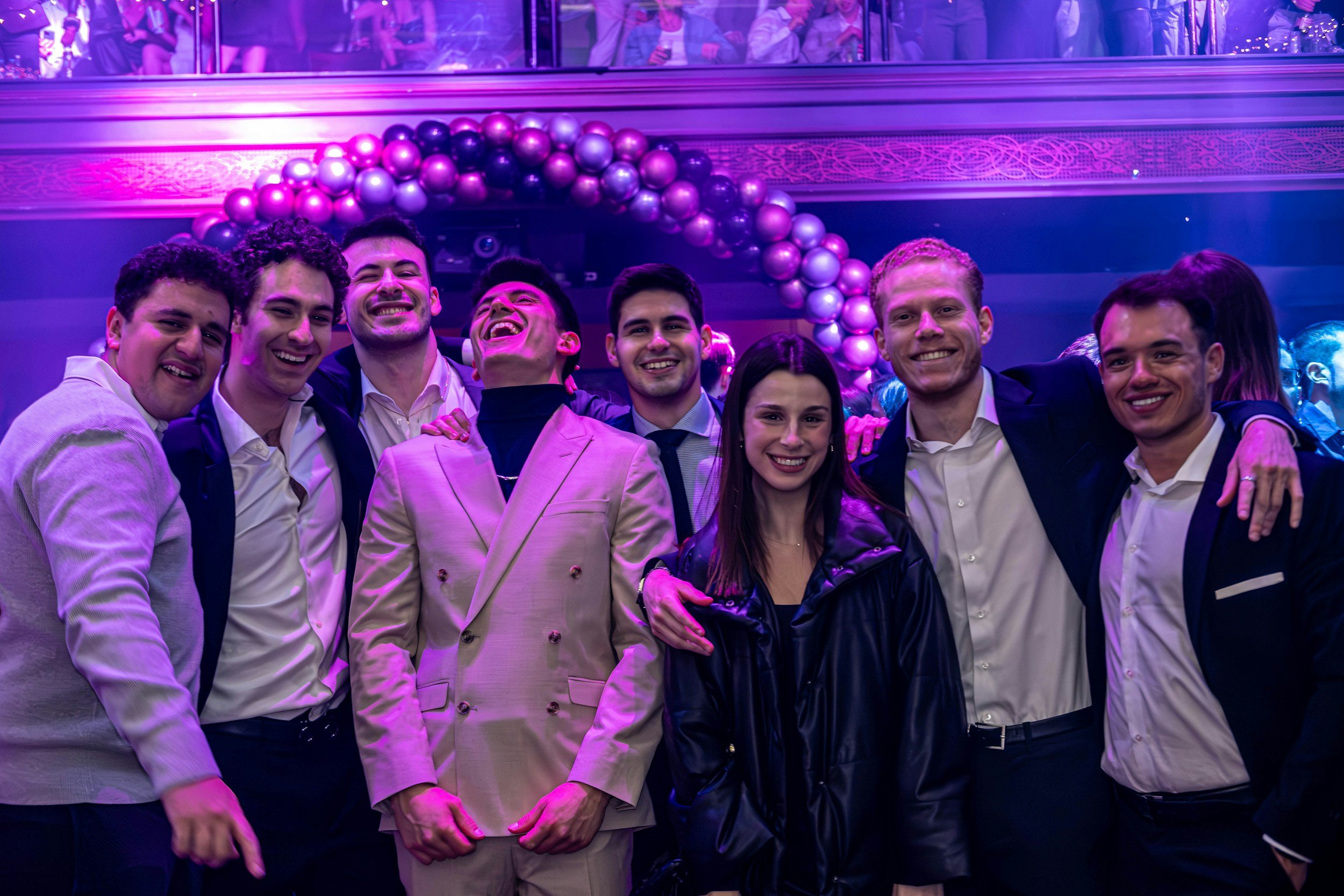 Group of people smiling and posing together at a lively party or celebration with purple and pink balloons and vibrant lighting.