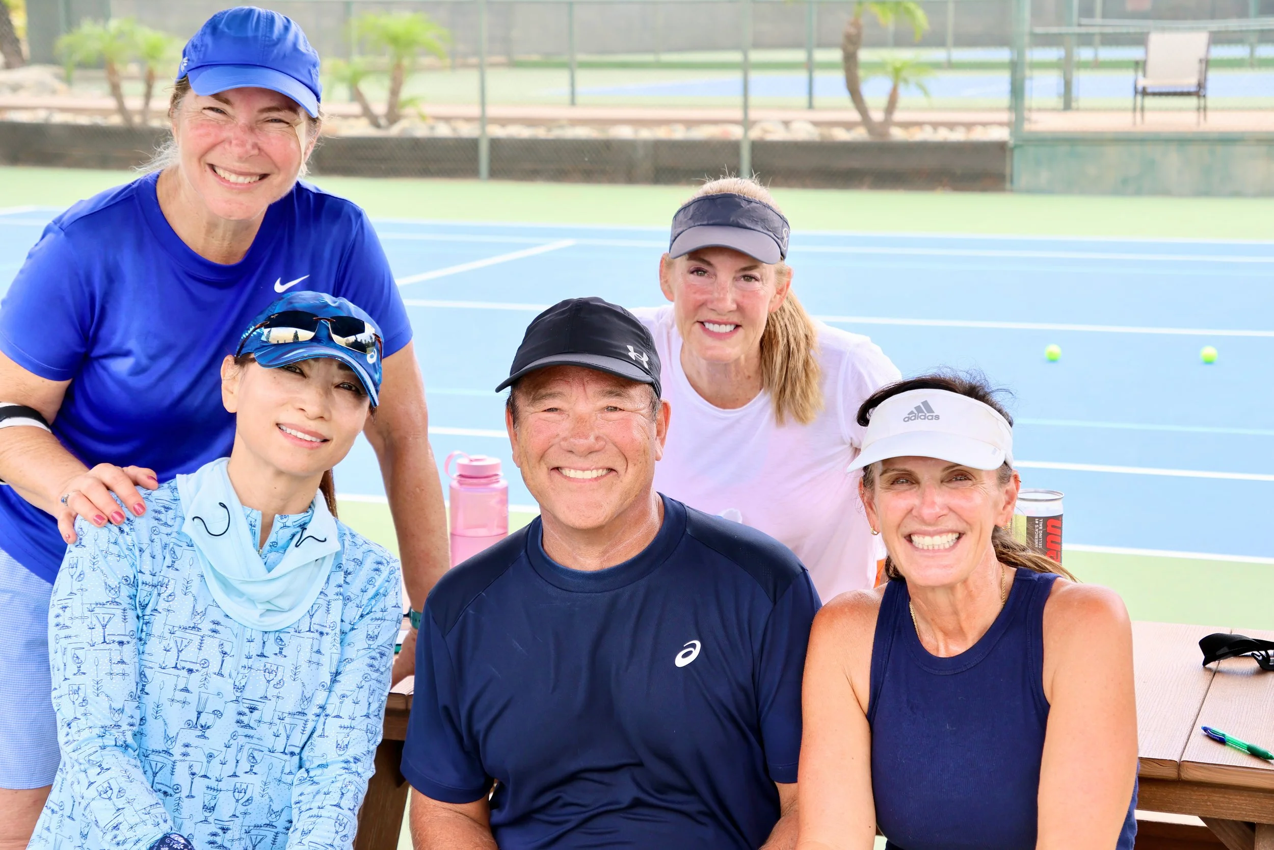 Group of six people smiling and posing for a photo on a tennis court, with tennis balls in the background, some wearing sports attire and hats.