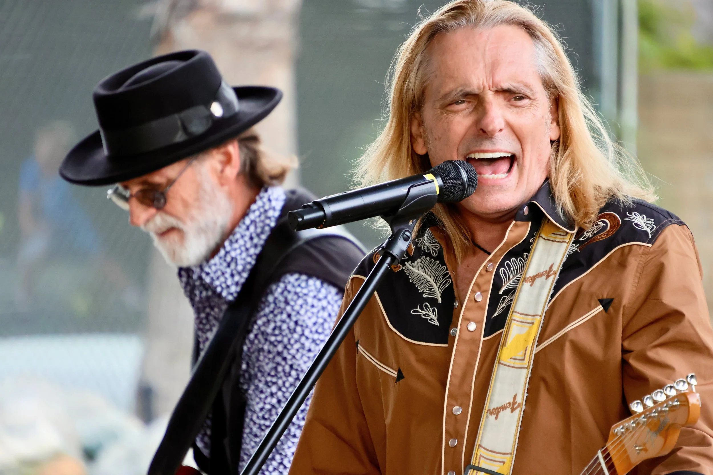 Two musicians performing outdoors, one with long blonde hair singing into a microphone and the other wearing a black hat and glasses.