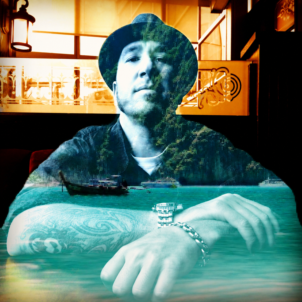 A double exposure photo combining a man wearing a hat and a black shirt with tattoos on his arm, sitting at a table with his arms crossed, and a scenic landscape of lush green mountains, water, and boats.