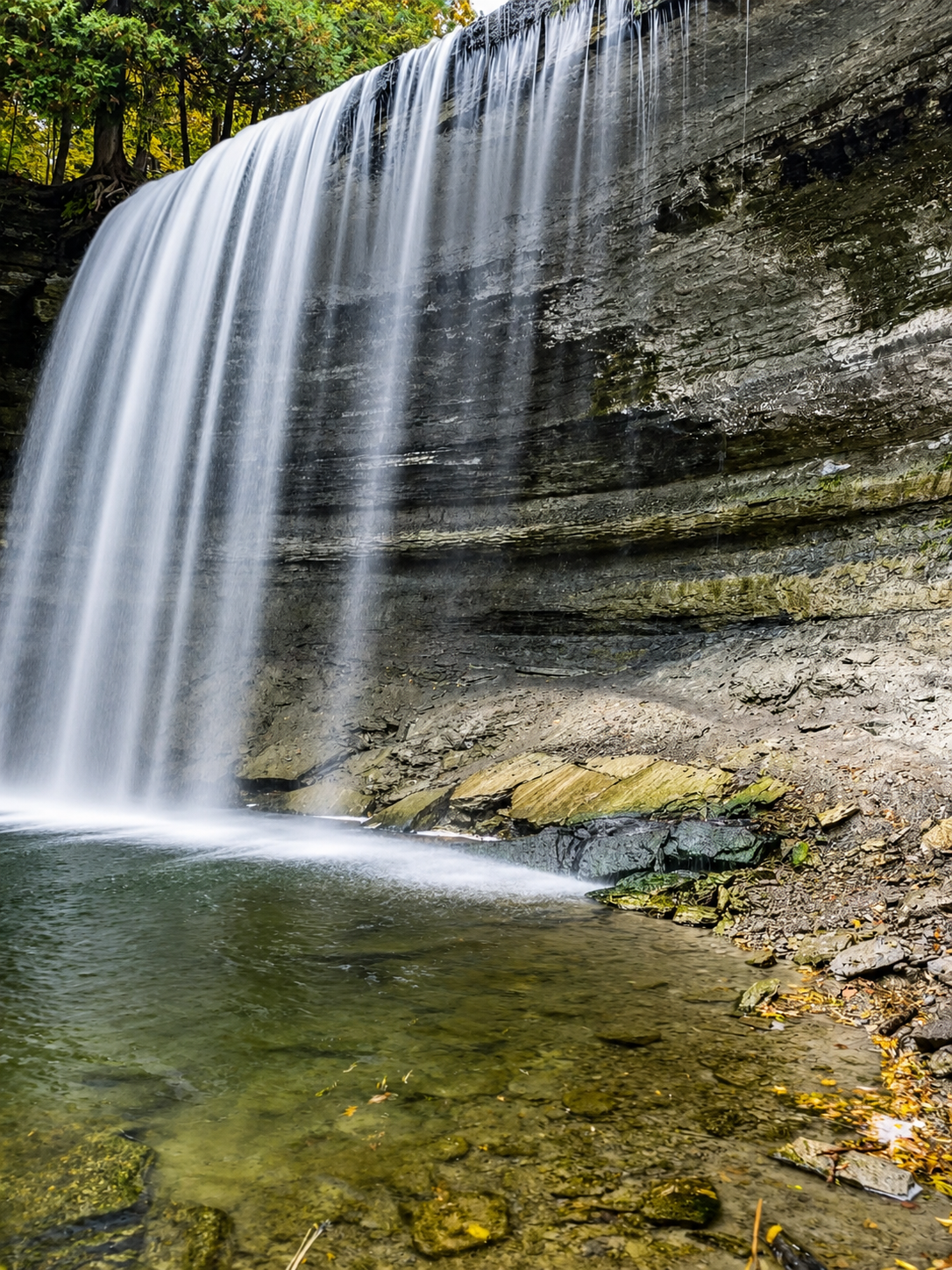 Waterfall over a rocky cliff with an emerald pool below and green trees above.