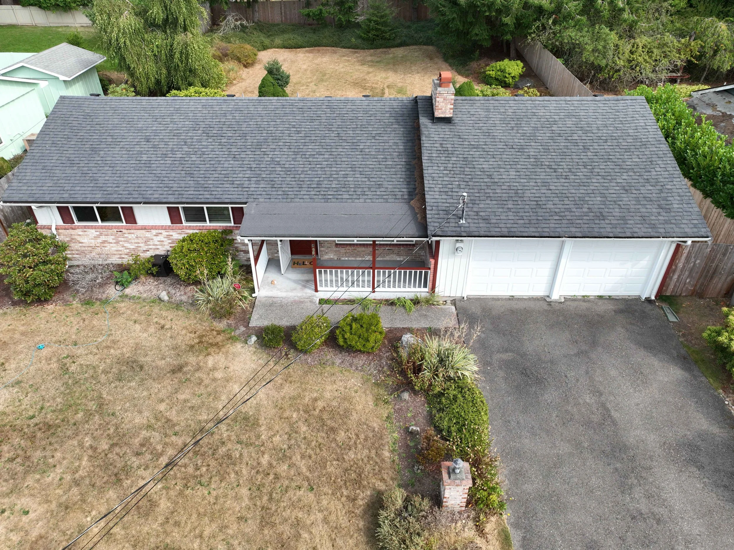 Aerial view of a single-story house with a gray shingled roof, a brick chimney, and a small front porch. The yard has a mix of grass, shrubs, and trees, with a fenced backyard that includes a lawn area and surrounding greenery.