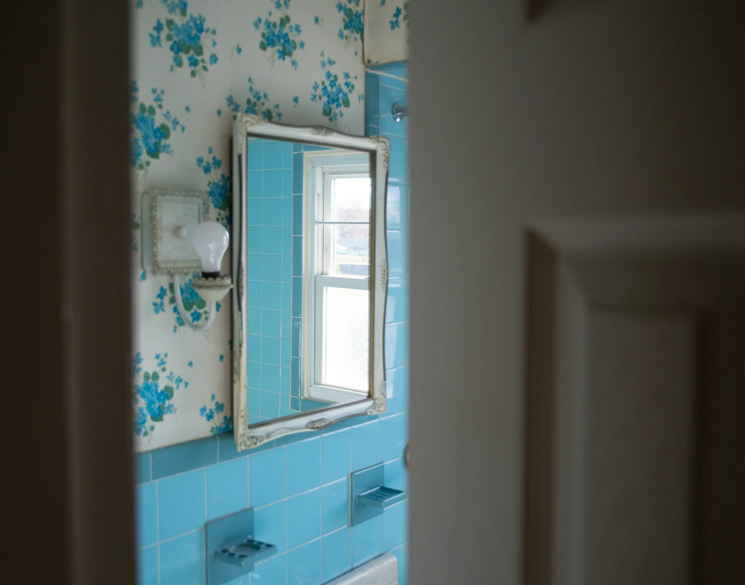 A bathroom mirror above a sink with a floral wallpaper background and a window in the background, viewed from a slightly tilted angle.