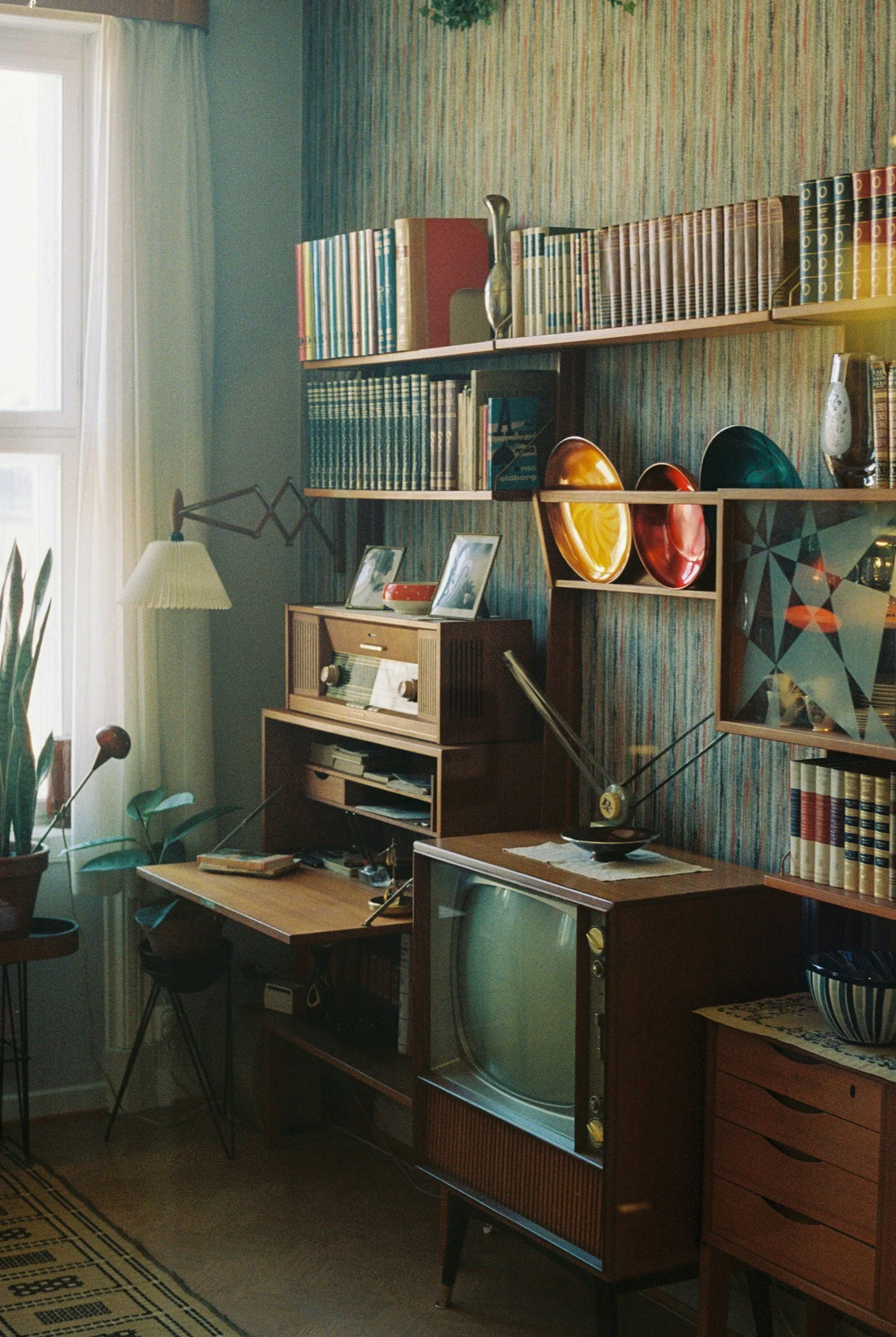 A vintage living room corner with shelves filled with books, a small wooden cabinet with a radio, a tin box, and picture frames, a wooden desk with an old TV, and a small table with a bowl, set against striped wallpaper and a window with curtains.