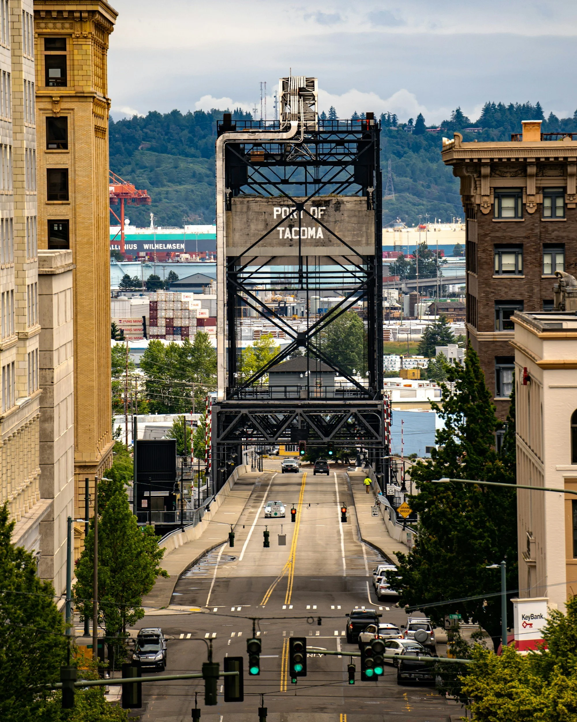 Downtown Tacoma city street with buildings on both sides, The Murray Morgan bridge, and a large ship in the water in the background; traffic lights are green.