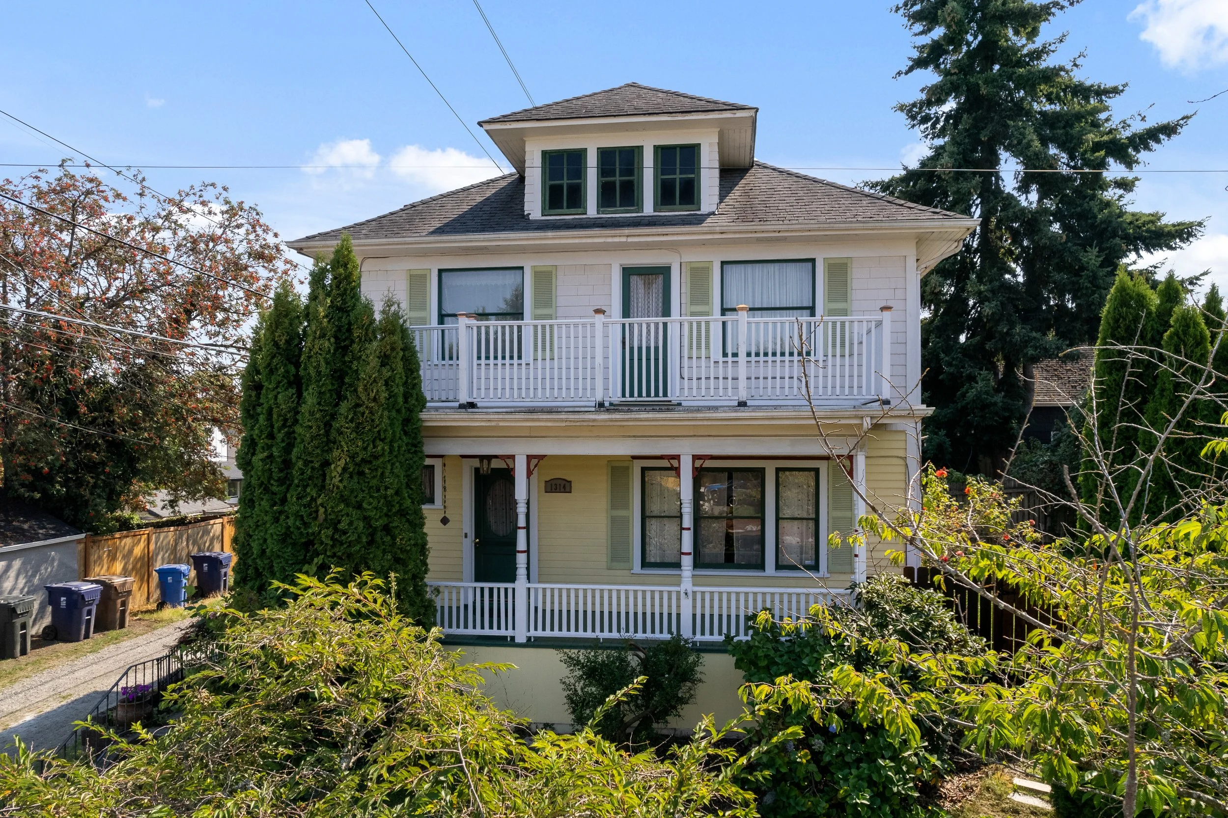 A three-story house with a gabled roof, white siding, and green window shutters, partially obscured by trees and bushes in a suburban neighborhood.