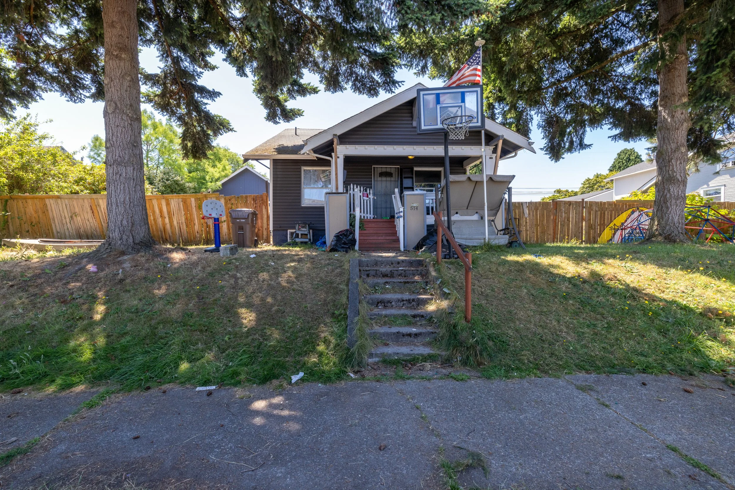 Backyard with step staircase leading to house, large trees, children’s basketball hoop, American flag, and playground equipment, wooden fence surrounding the yard.