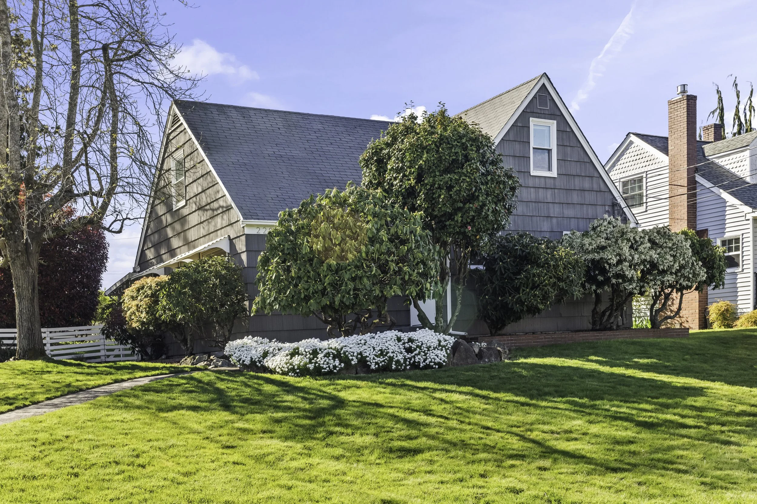 Front yard of a house with a manicured lawn, colorful flower beds, trees, and shrubs, under a clear blue sky.