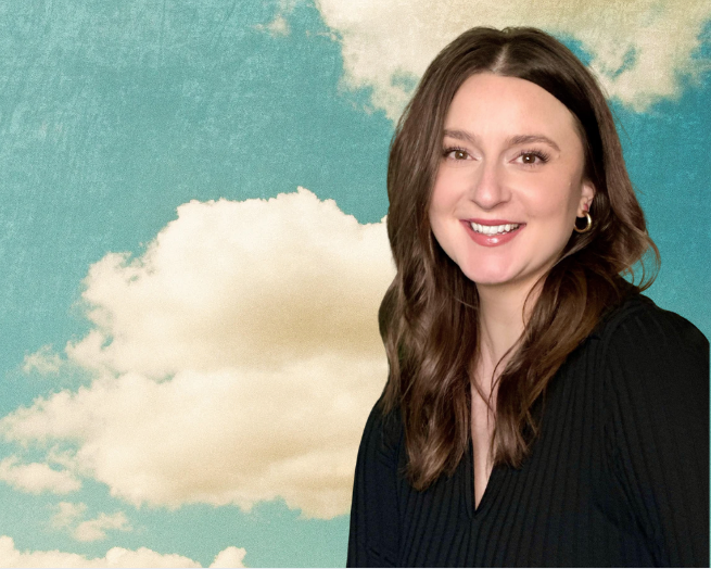 Lora- Root Real Estate Broker smiling, standing in front of a sky background with clouds.