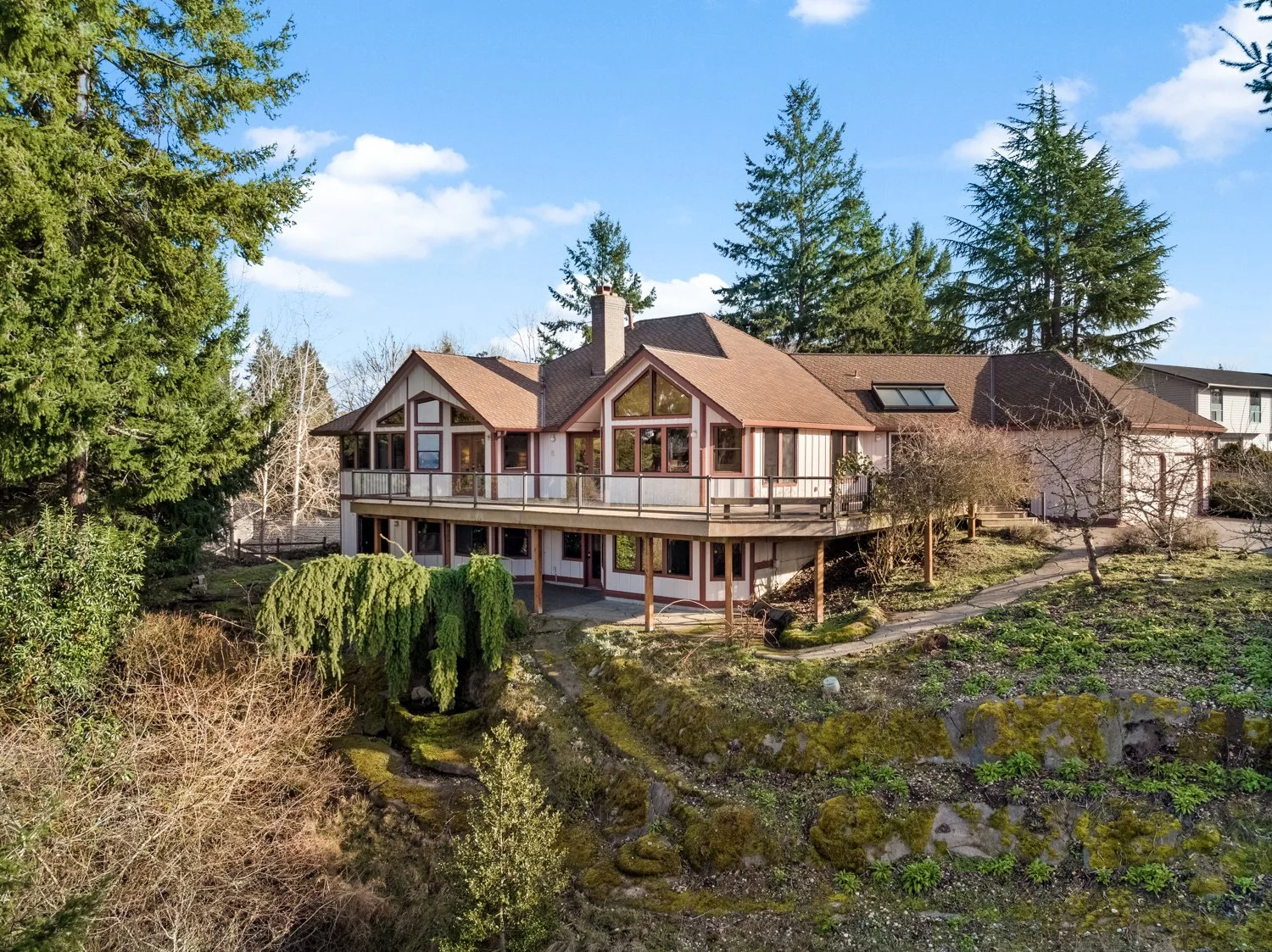 A house with large windows and a wooden balcony, surrounded by tall trees and a garden with rocks and small plants, under a blue sky with some clouds.