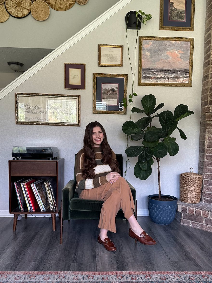 A woman with long, wavy brown hair sitting on a dark green armchair, smiling. The background features a white wall decorated with framed paintings and pictures, a large potted fiddle leaf fig plant, a wicker basket, a small wooden record player stand with records, and a brick fireplace on the right.