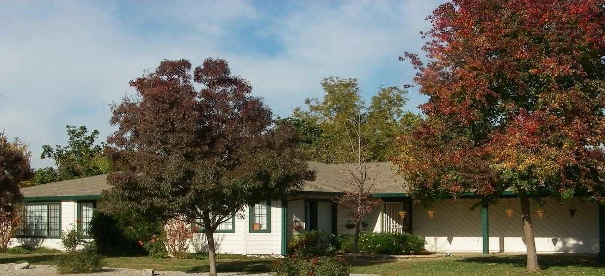 A white house with a green roof surrounded by trees with fall foliage in red, orange, and green, under a partly cloudy blue sky.