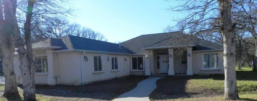 Single-story house with a front porch, surrounding trees, and a curved walkway leading to the front door.