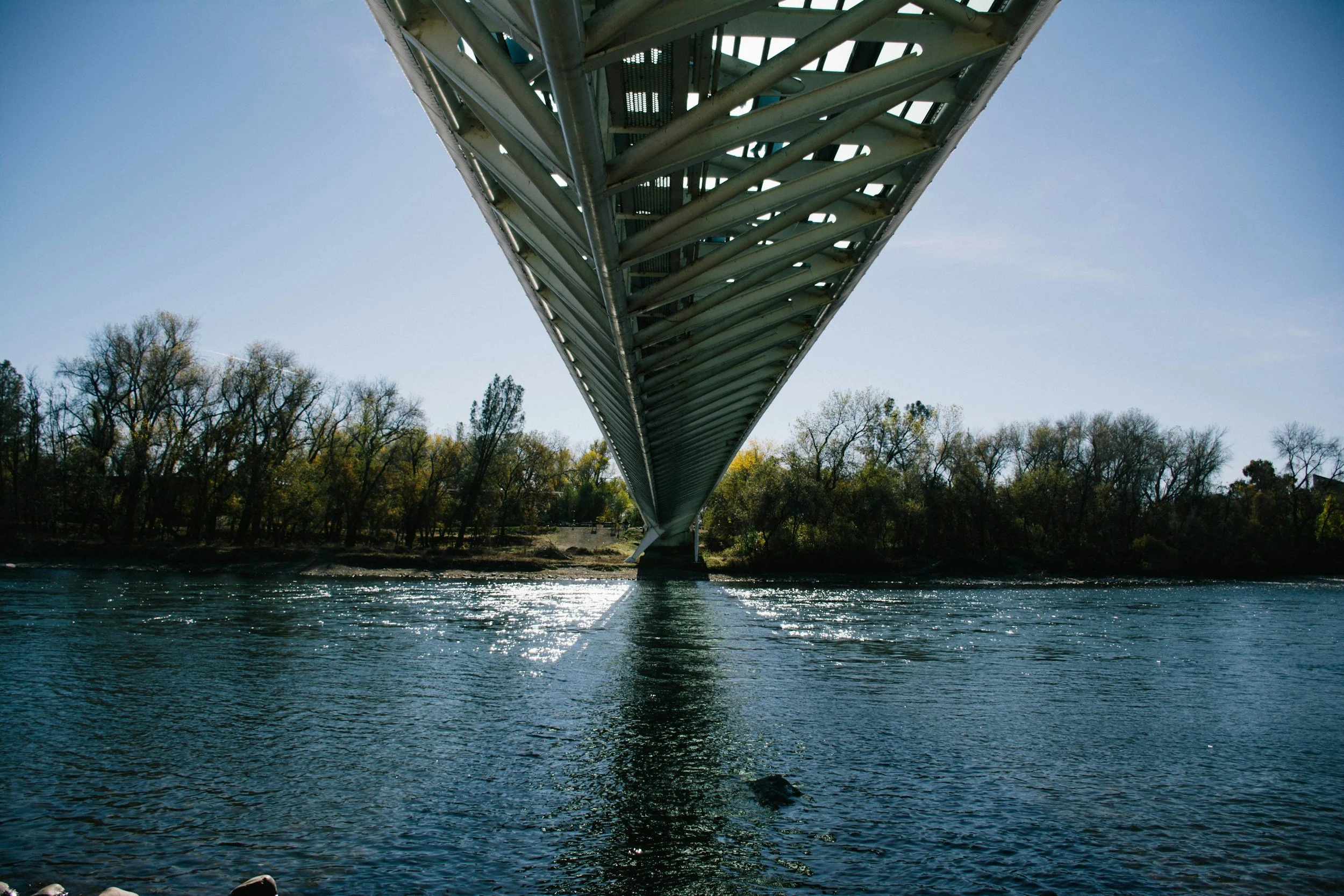 Looking up at the underside of a large metal bridge spanning over a body of water, with trees in the background and sunlight reflecting on the water.