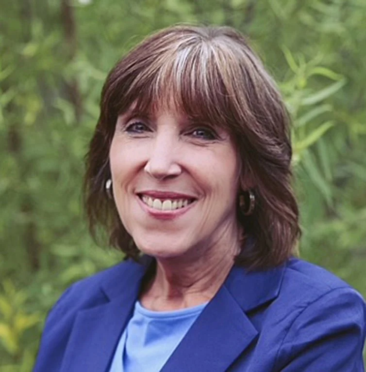 A woman with shoulder-length brown hair wearing a blue blazer and a light blue top, smiling outdoors with green foliage in the background.