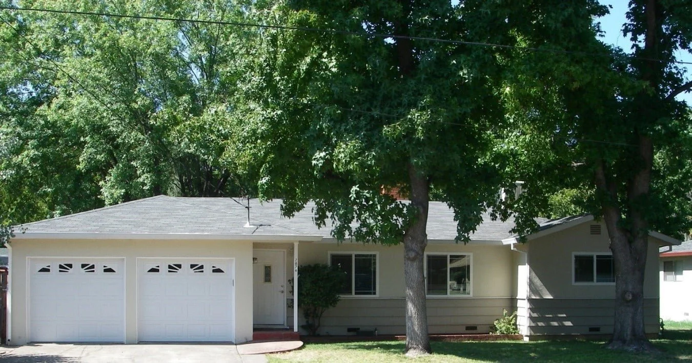 Front view of a single-story house with two garage doors, surrounded by large trees with green foliage.