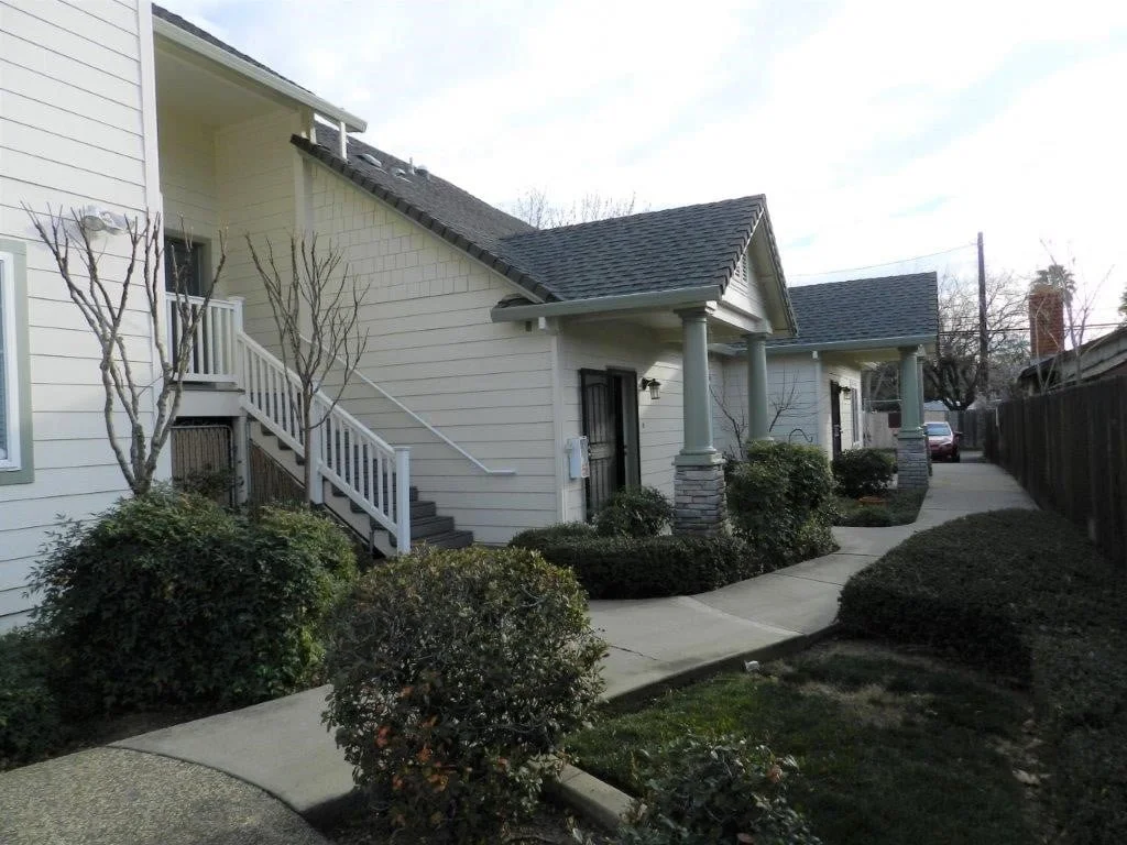 Exterior view of a modern house with beige siding, black roof, and a sidewalk leading to the front door. The house features white columns, a staircase, leafless trees, bushes, and a wooden fenced yard.