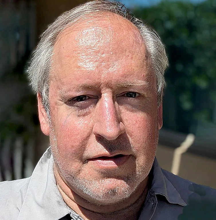 Close-up photo of a middle-aged man with light skin, gray hair, and a slight beard, outdoors with a blurred green background.