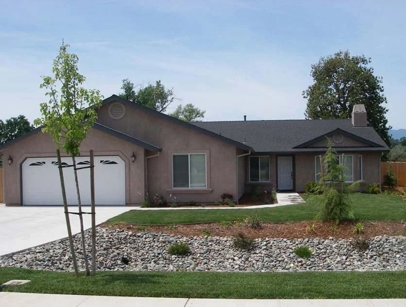 A single-story house with a beige exterior, black roof, and a front yard featuring a grass lawn, small trees, and decorative rocks.