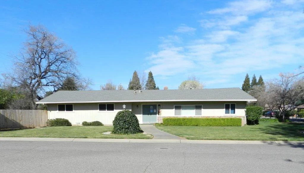 Single-story beige house with a gray roof, front yard with trimmed bushes, concrete pathway, and trees on a sunny day.